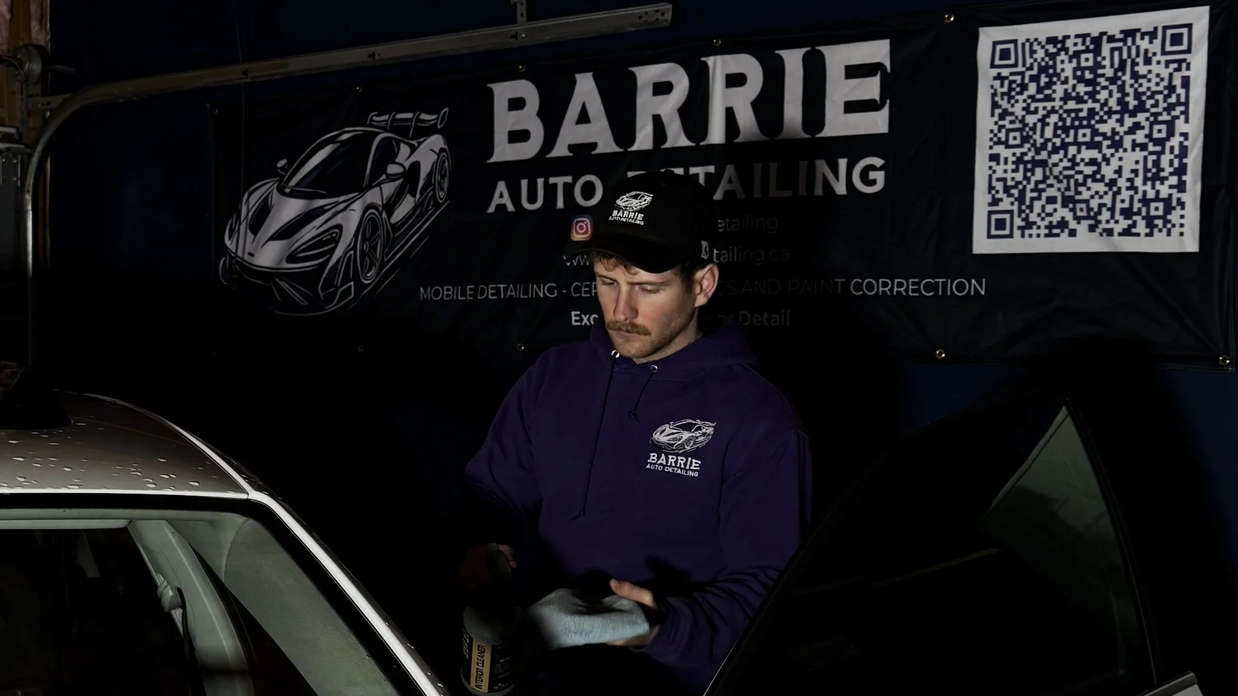 A man wearing a Barrie Auto Detailing hoodie and cap working on a car in a garage with a Barrie Auto Detailing banner in the background.