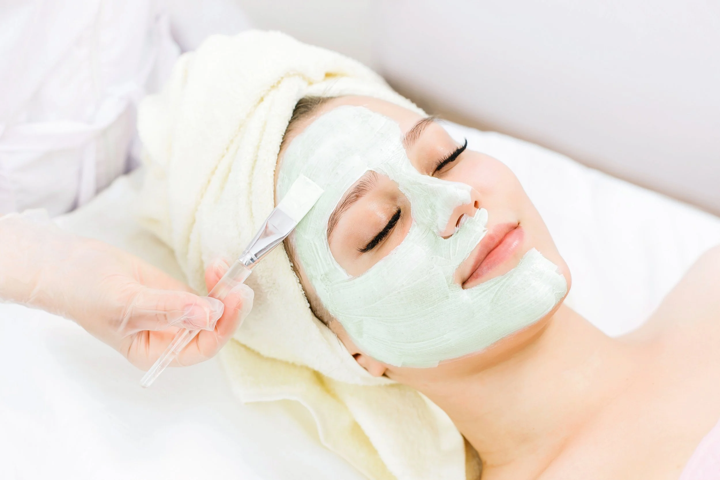 Bioline Primaluce Brightening: Woman lying down with eyes closed, receiving a facial treatment with green mask applied to her face, wrapped in a towel, in a spa or skincare clinic.