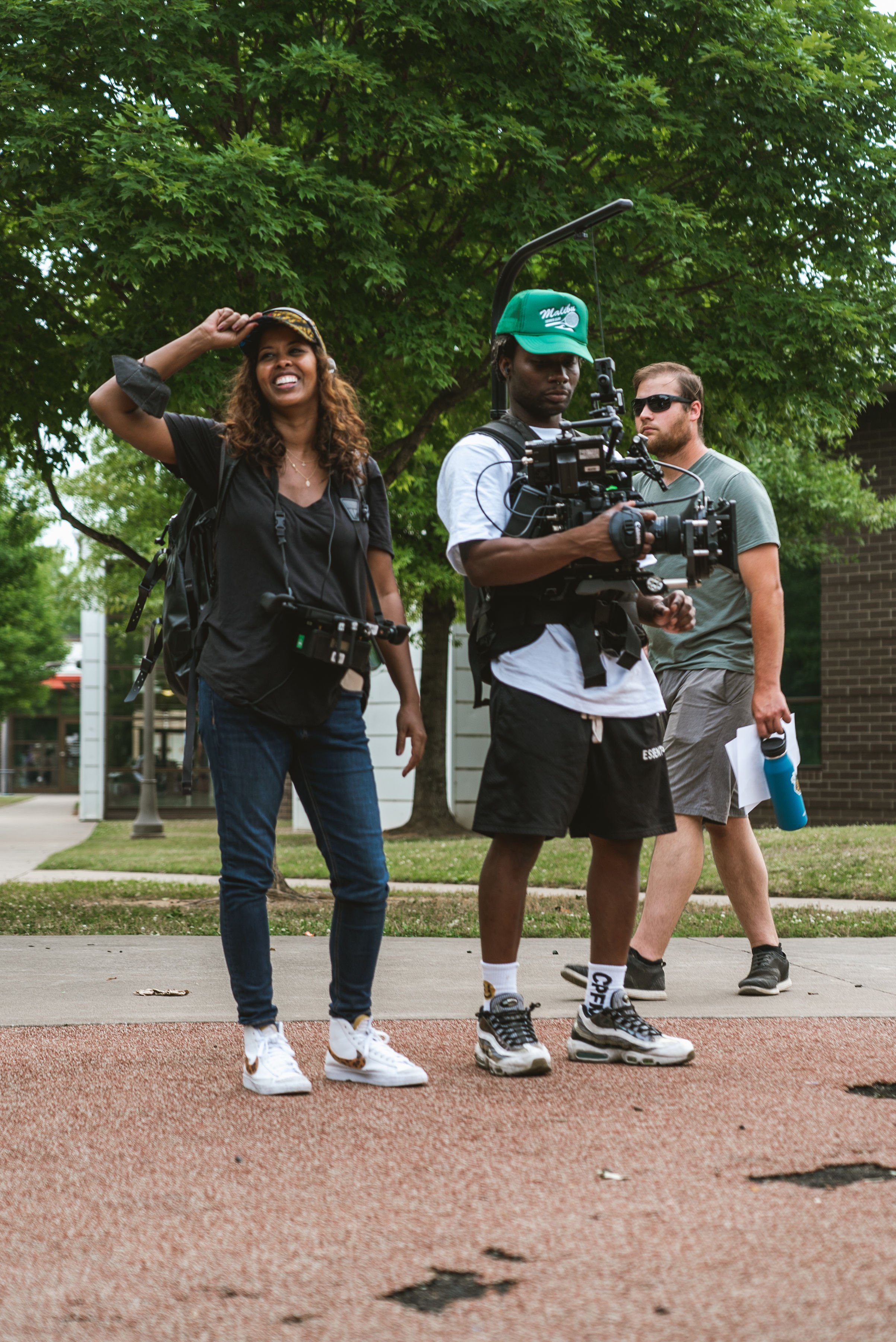 idil ibrahim and Fletch power ; one carrying professional camera equipment, another smiling and adjusting a cap, with greenery and buildings in the background.