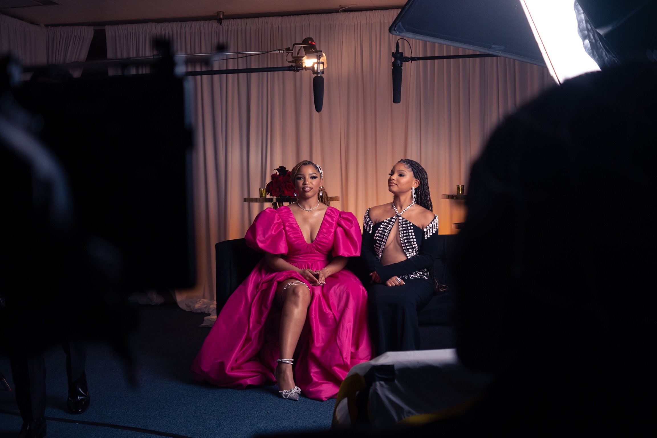 Two women seated in a studio setting, one wearing a bright pink dress and the other in a black dress, with studio lights and microphones visible.