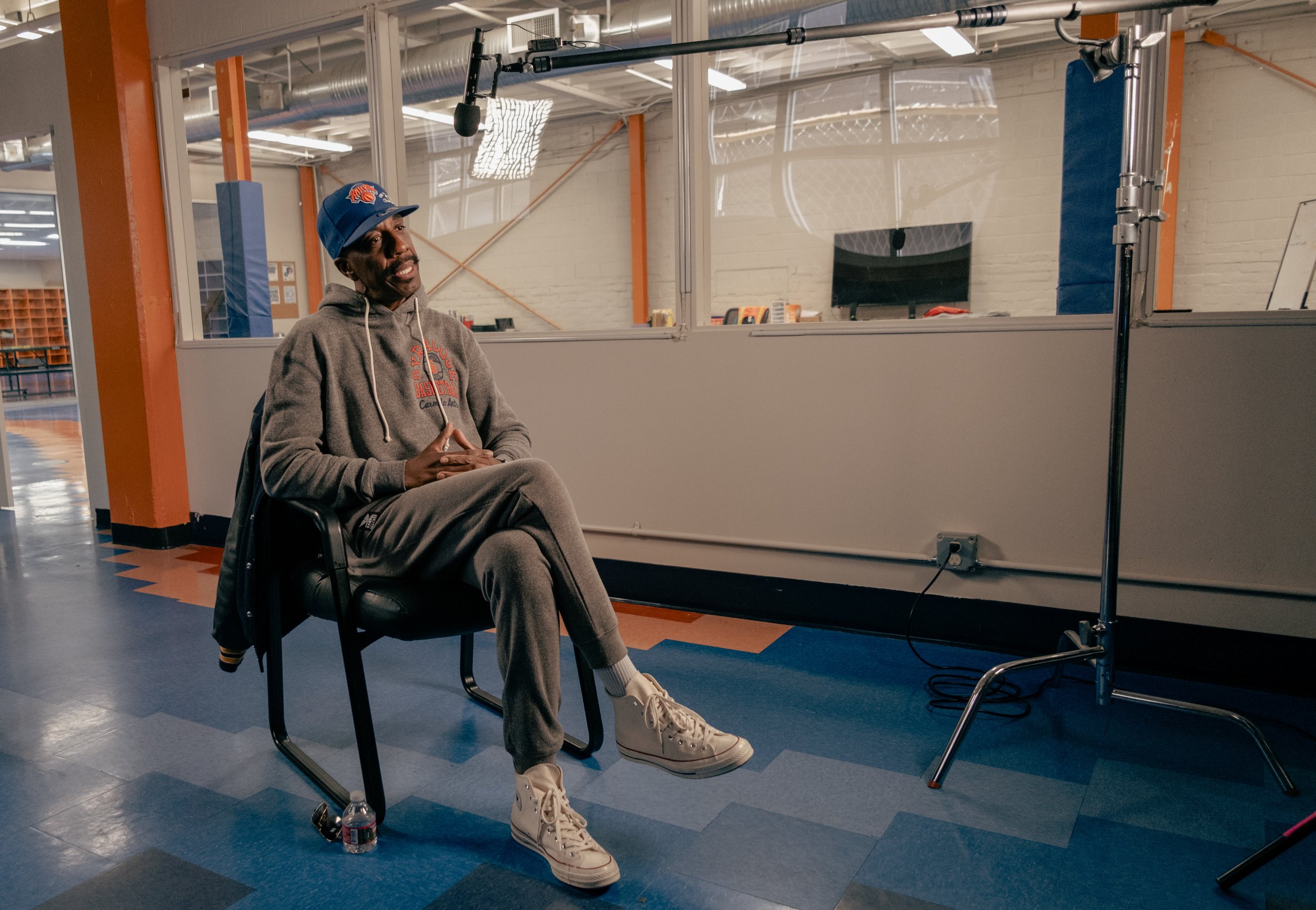 Person wearing a gray tracksuit and a blue cap, sitting in a chair with a microphone overhead in a studio setting.