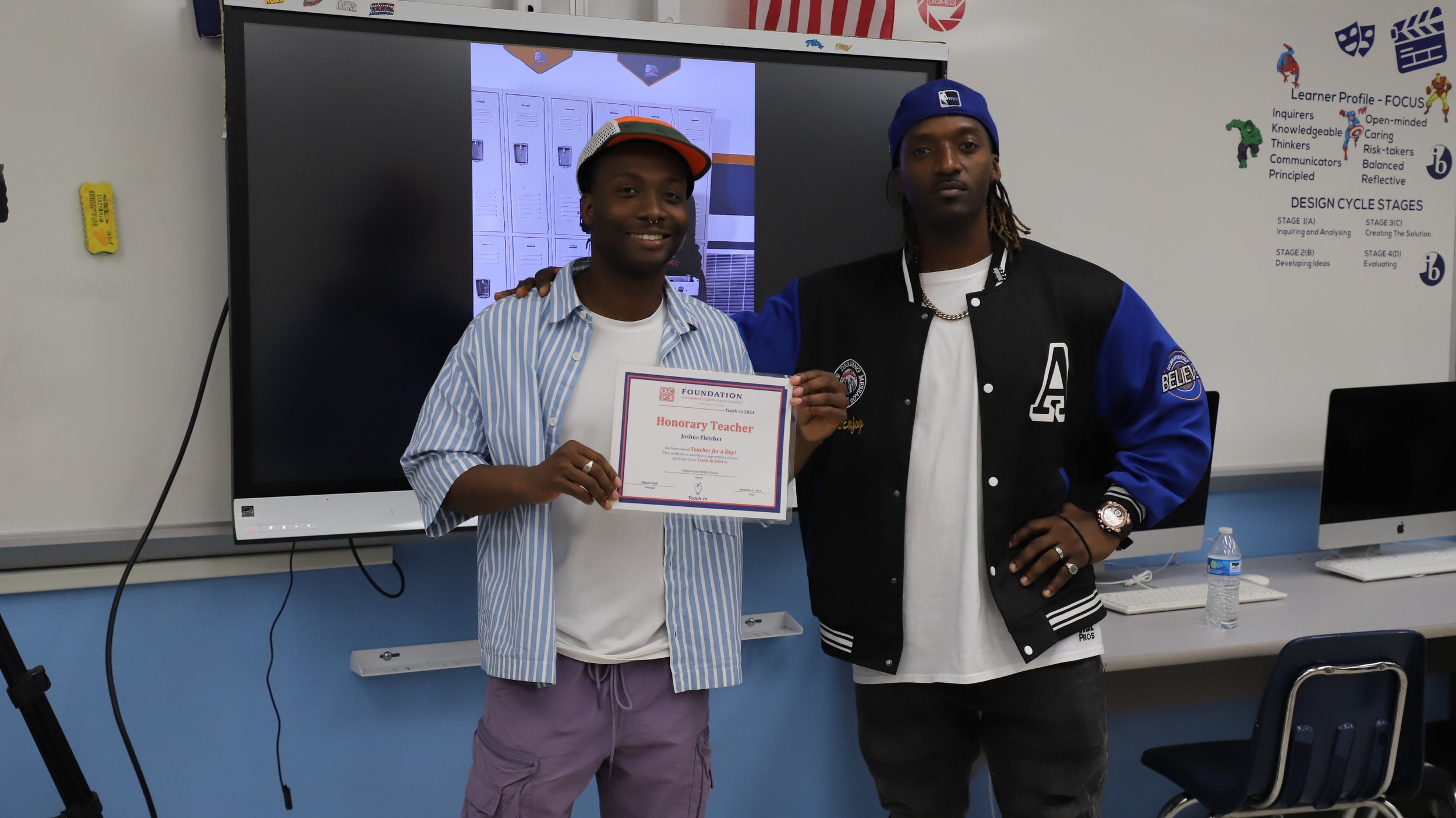 Fletch Power posing in a classroom, holding an "Honorary Teacher" certificate, standing in front of a large screen and computers.