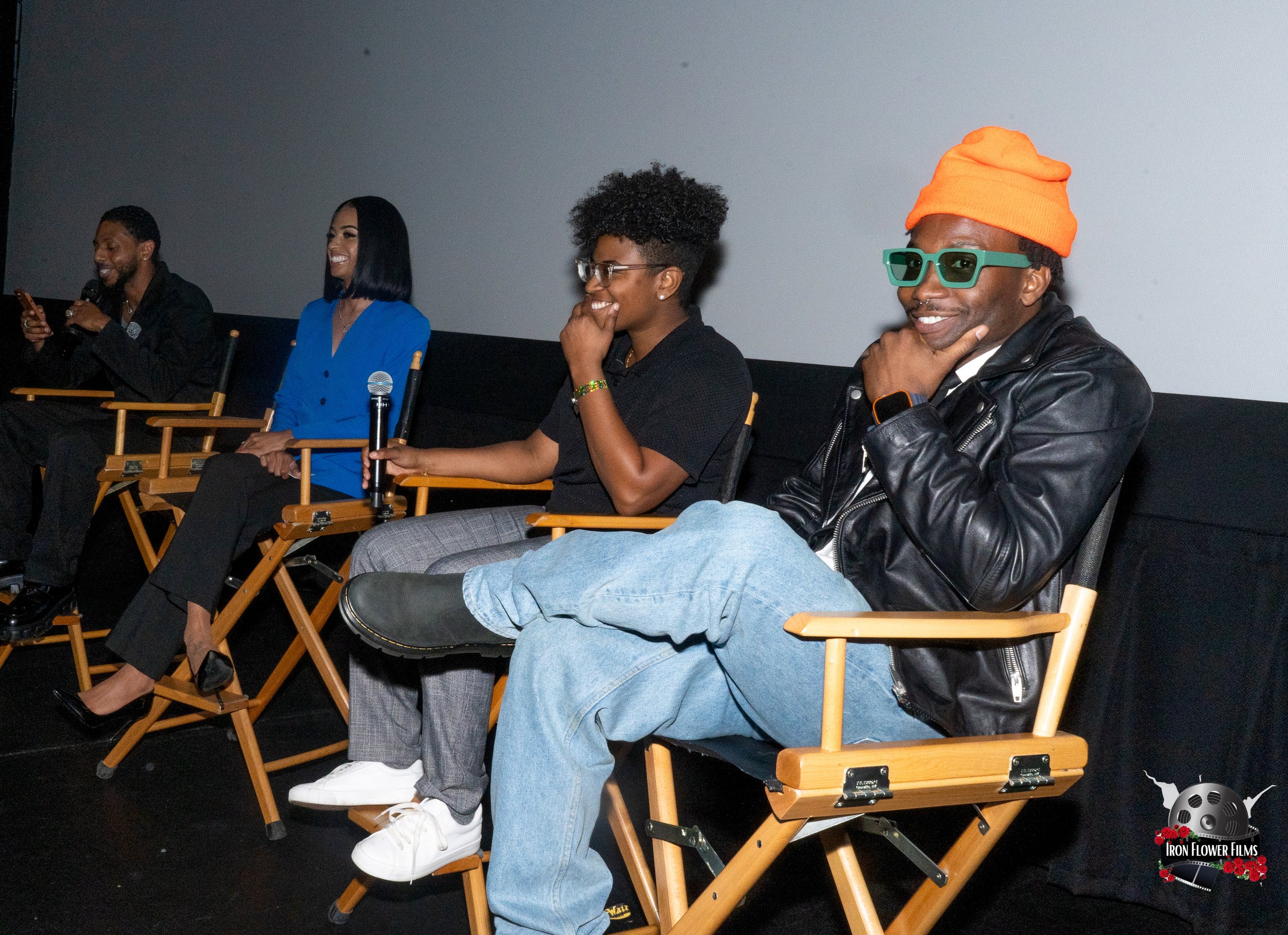 fletch power and a Group of people sitting on director's chairs at a panel discussion in a theater, holding microphones, with a simple stage and projection screen in the background. Fletch wears an orange beanie and sunglasses.