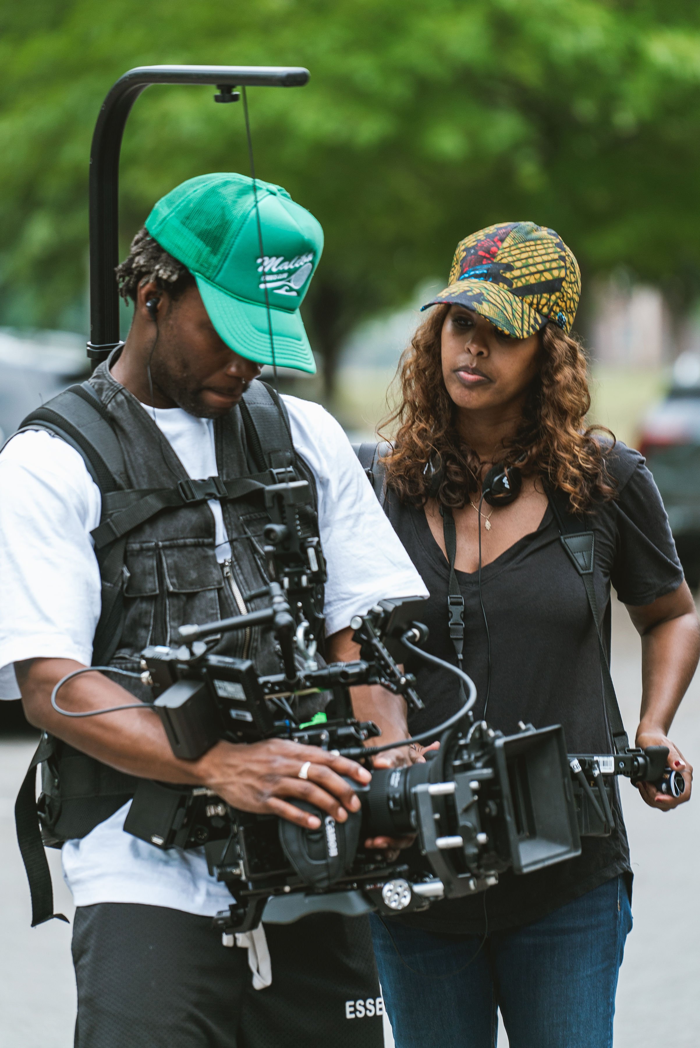 Two people on a film set, one operating a professional camera with a stabilizer and the other observing. Both are wearing casual clothing and hats, with the person on the right having headphones around their neck. Background is blurred with greenery.
