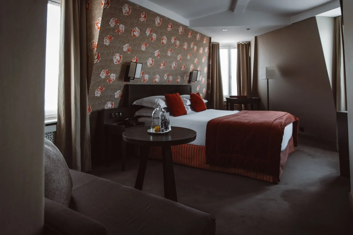 Chambre d'hôtel élégante décorée avec du papier peint floral rouge, lit double avec couette rouge et oreillers assortis, table ronde, bouquet sous cloche, lumières murales, et fenêtres avec rideaux beiges.