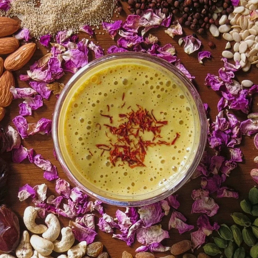 A glass of golden-yellow beverage garnished with saffron strands, surrounded by scattered dried rose petals, almonds, cashews, dried berries, pumpkin seeds, and other assorted nuts and dried fruits on a wooden surface for ayurvedic postpartum.