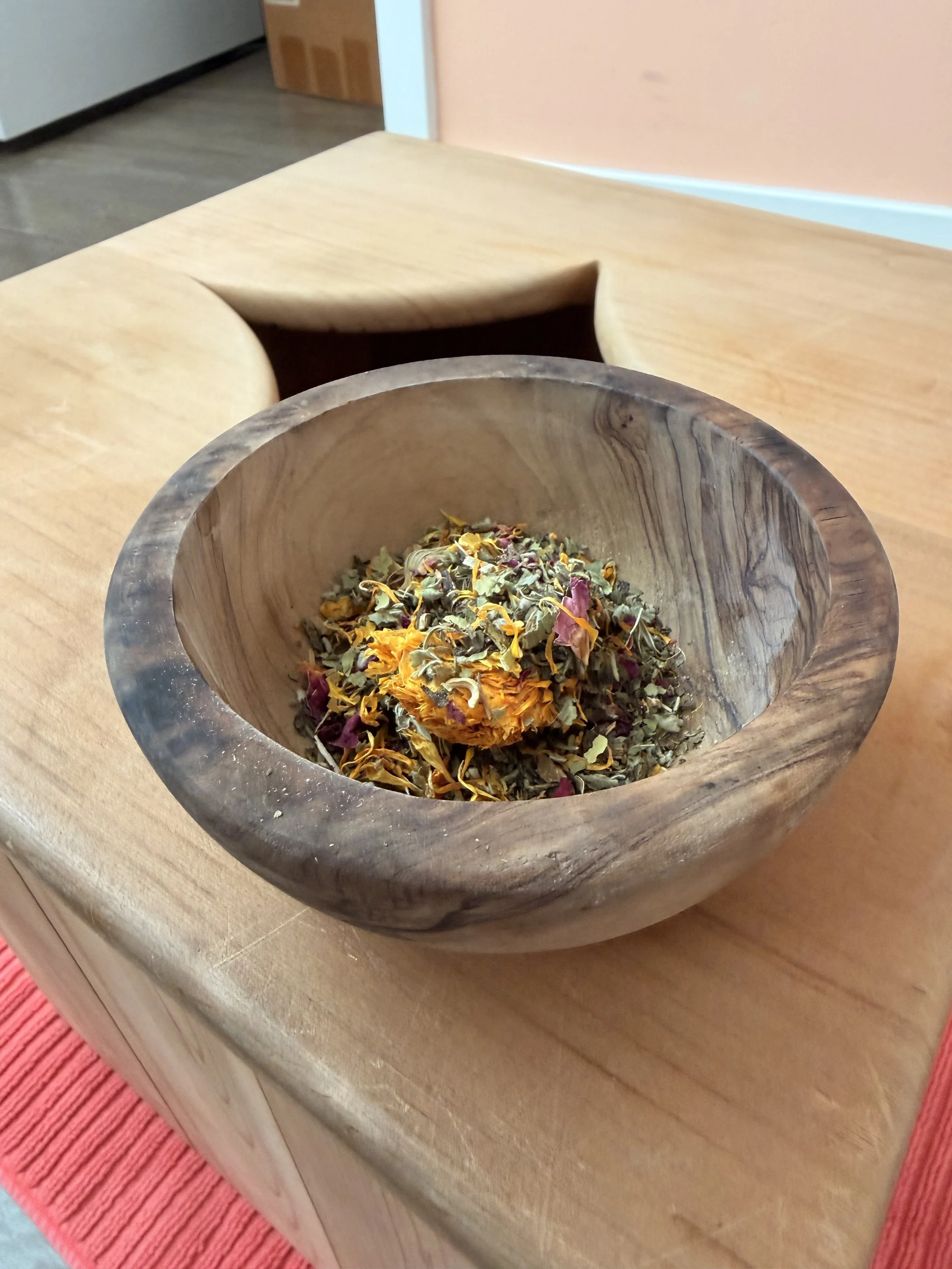 A wooden bowl filled with dried herbs and flower petals placed on a wooden table.