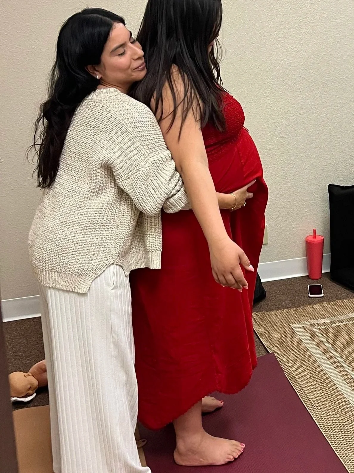 Two women hugging, one with long dark hair in a cream sweater and white striped pants, and another in a red dress with long dark hair, standing barefoot on a yoga mat.