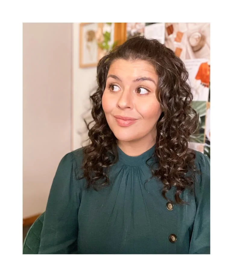 Chloe Forbes-Kindlen with hair down, brown curls and wearing a green dress looking at the text displayed on the right. Background shows custom designed wall collage