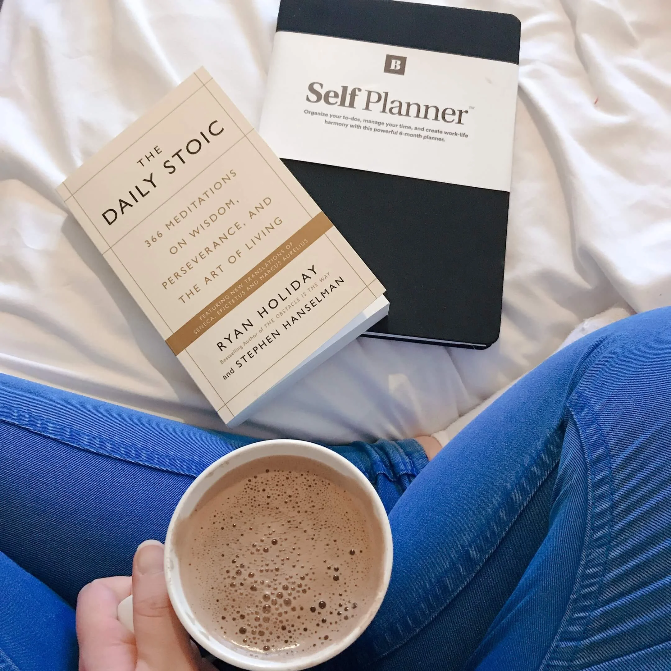 Chloe Forbes-Kindlen sitting cross-legged wearing jeans with cup of coffee in hand, the daily stoic book to the left and a best self self-planner to the right.