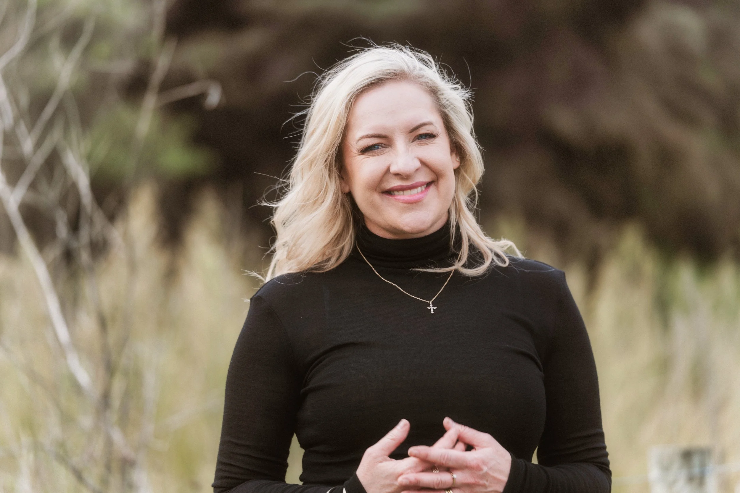 A woman with blonde hair wearing a black turtleneck, smiling outdoors with trees blurred in the background.
