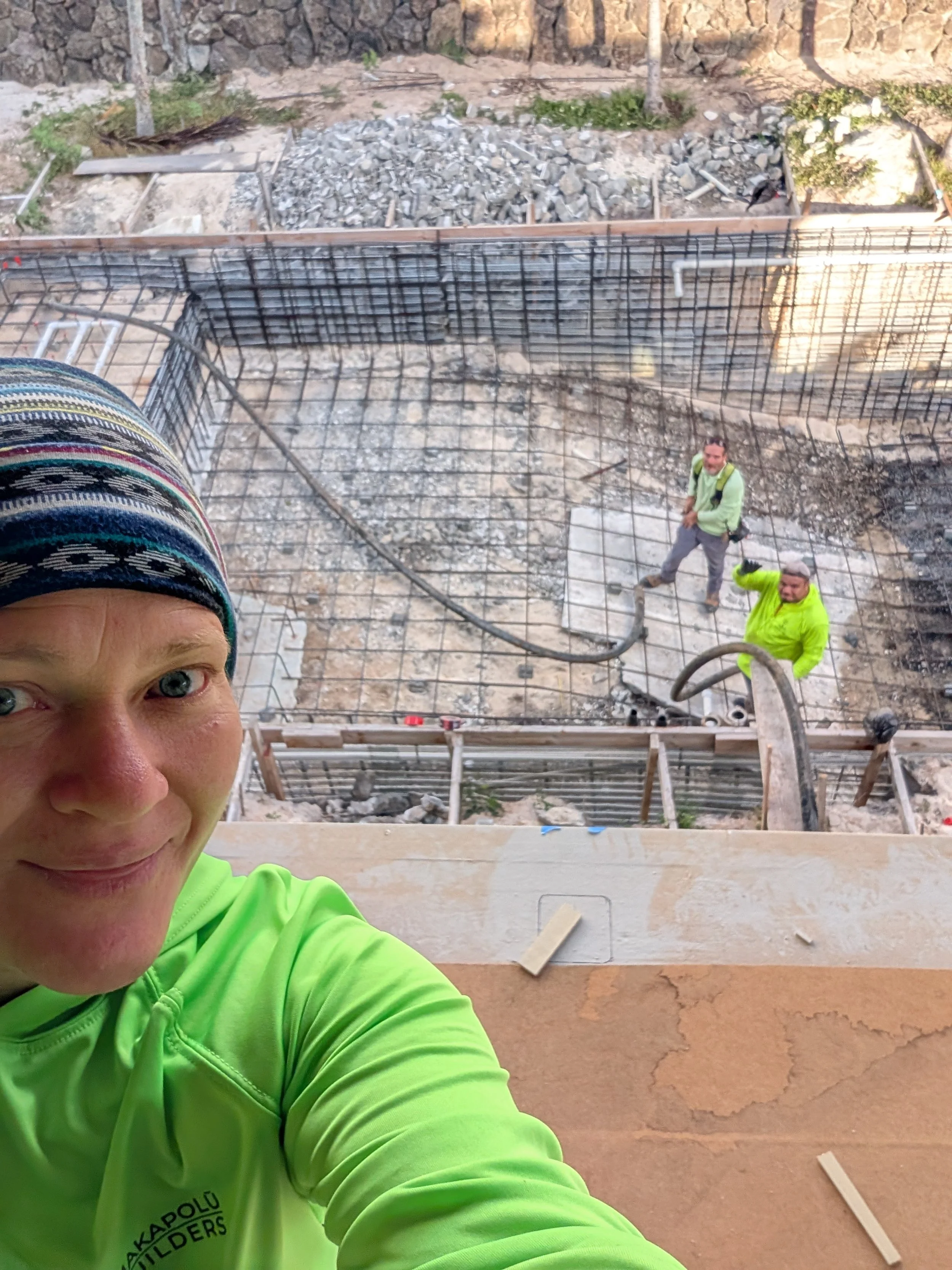 A person taking a selfie on a construction site, with two workers visible in the background working on the foundation, wearing safety gear, with rebar and concrete foundation visible.