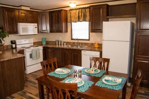 Kitchen with wooden cabinets featuring a white refrigerator, stove, and microwave. A brown dining table with four chairs is set with plates and turquoise placemats. The kitchen has a window and wood flooring.