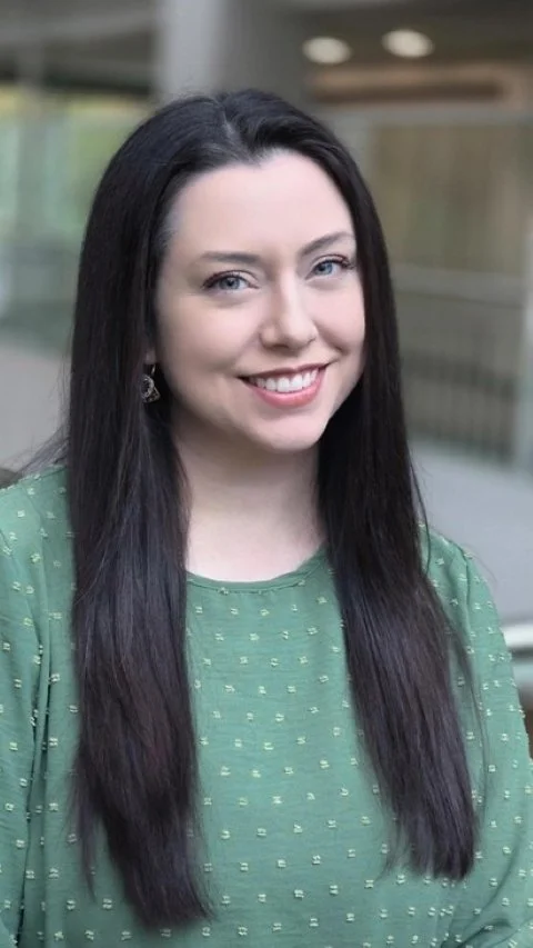 Portrait of a smiling woman with long dark hair, wearing a green top with small pattern, standing outdoors in an urban setting.