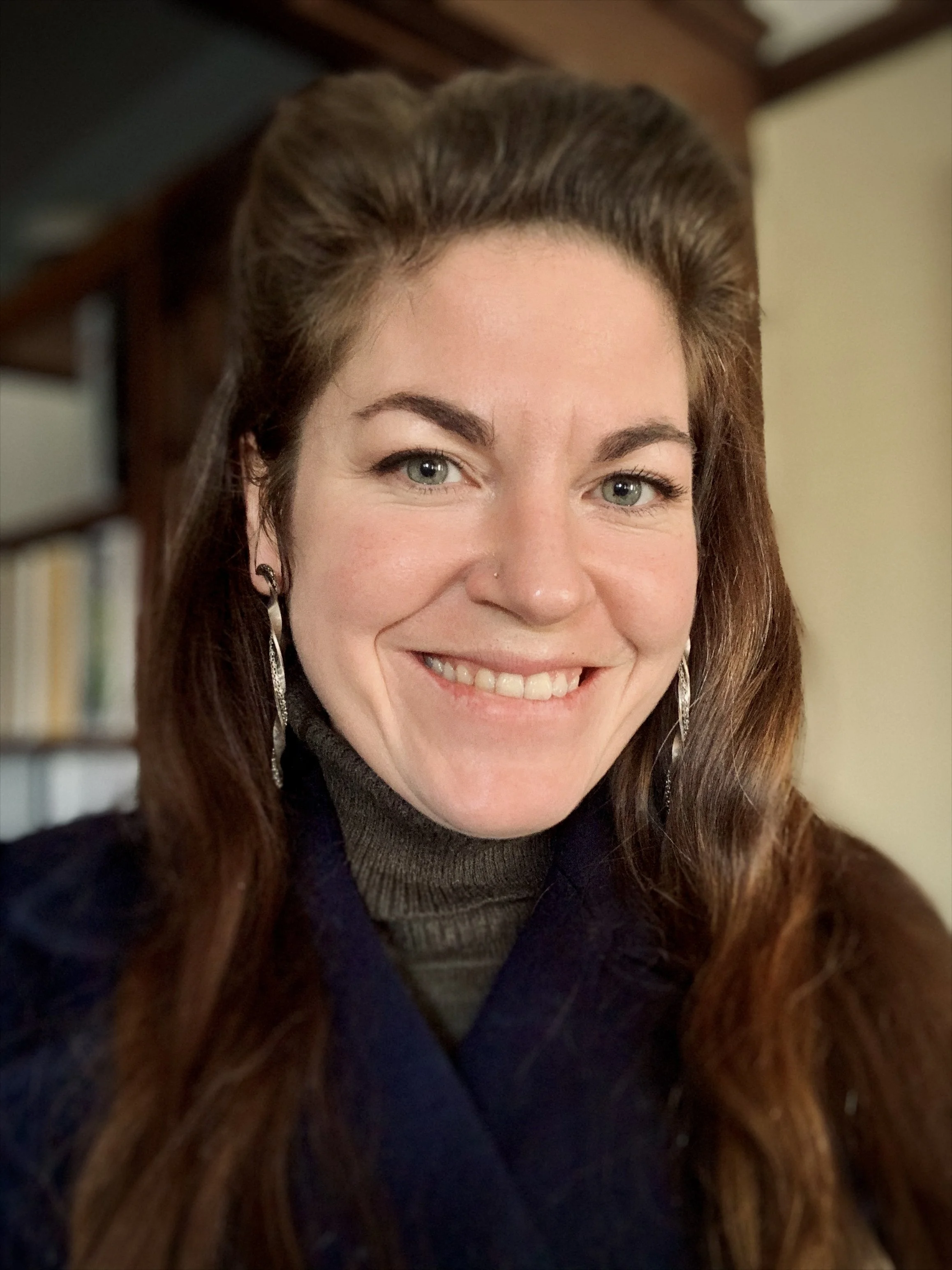 Close-up of a woman with long auburn hair, smiling, wearing a dark sweater and earrings, with bookshelves in the background.