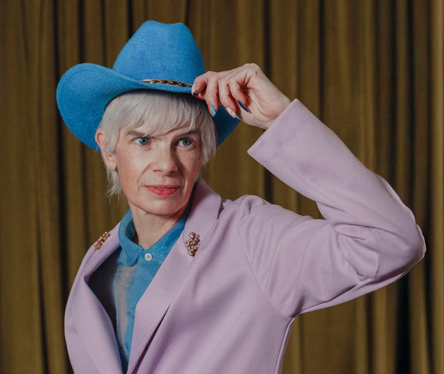 Rae looks sharp in a pink jacket and blue cowboy hat. They are smiling.