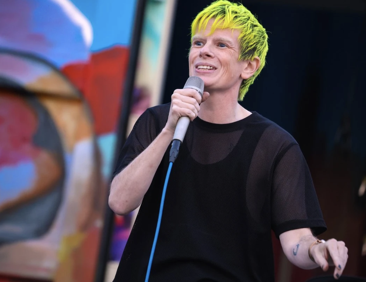 Rae sings into a microphone - they have bright yellow hair and a smile.