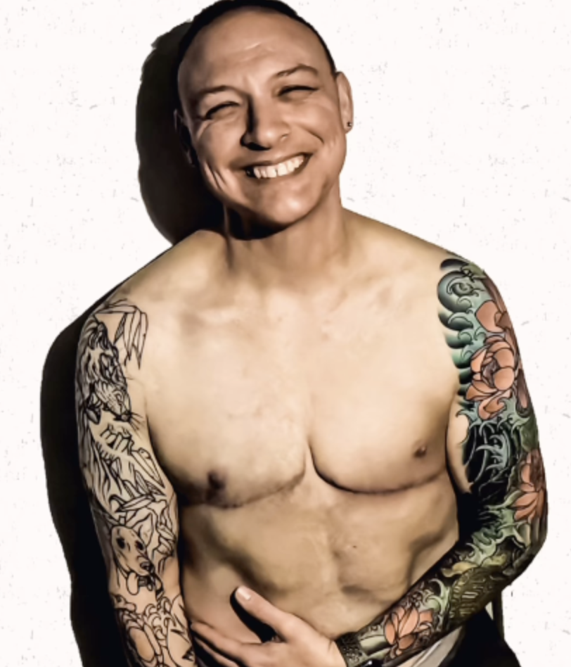Ash is a smiling trans man with colourful tattoos.