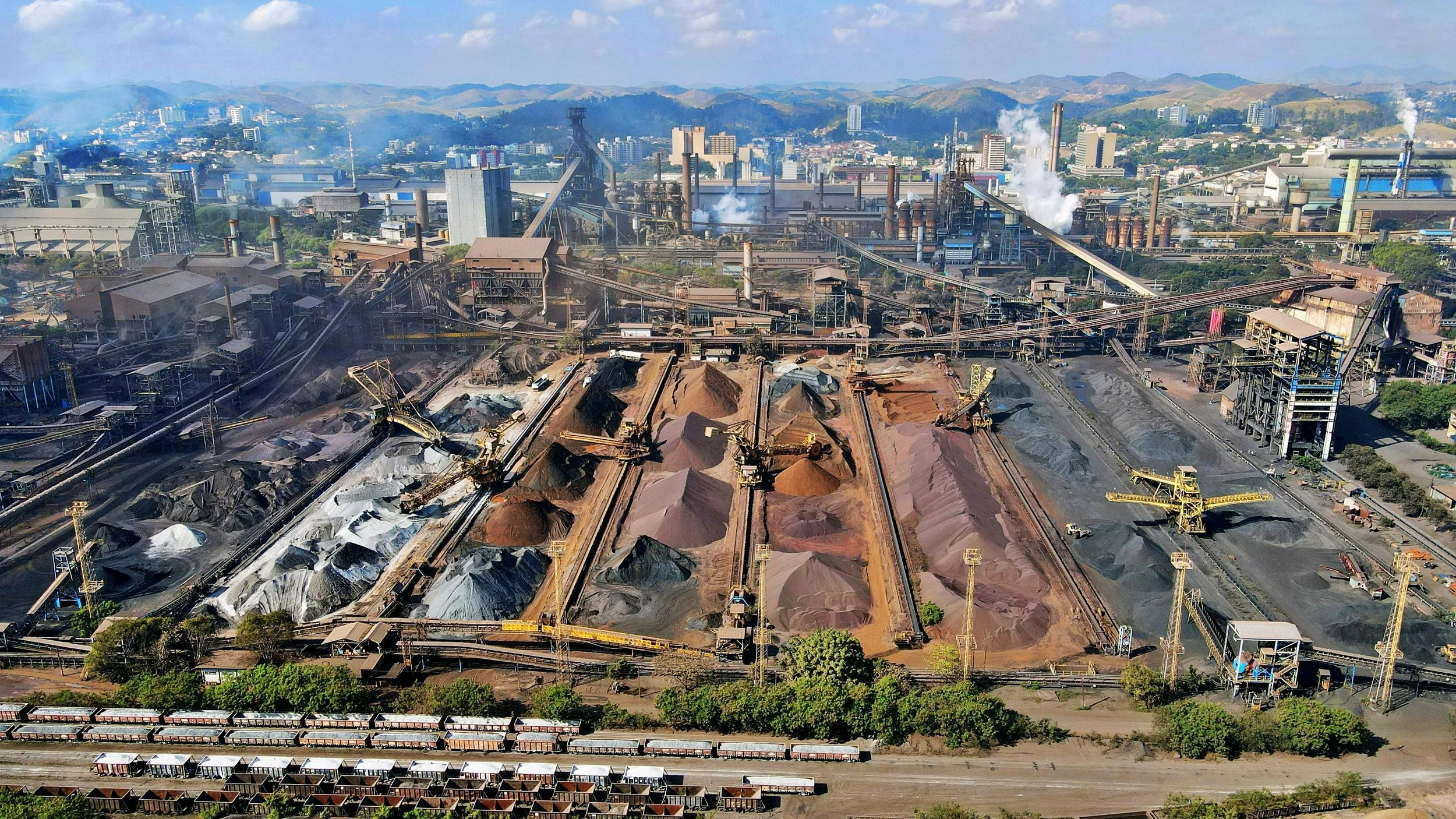 Vista aérea de una planta industrial con montículos de minerales de colores y maquinaria de excavación en primer plano, y una ciudad con montañas al fondo.