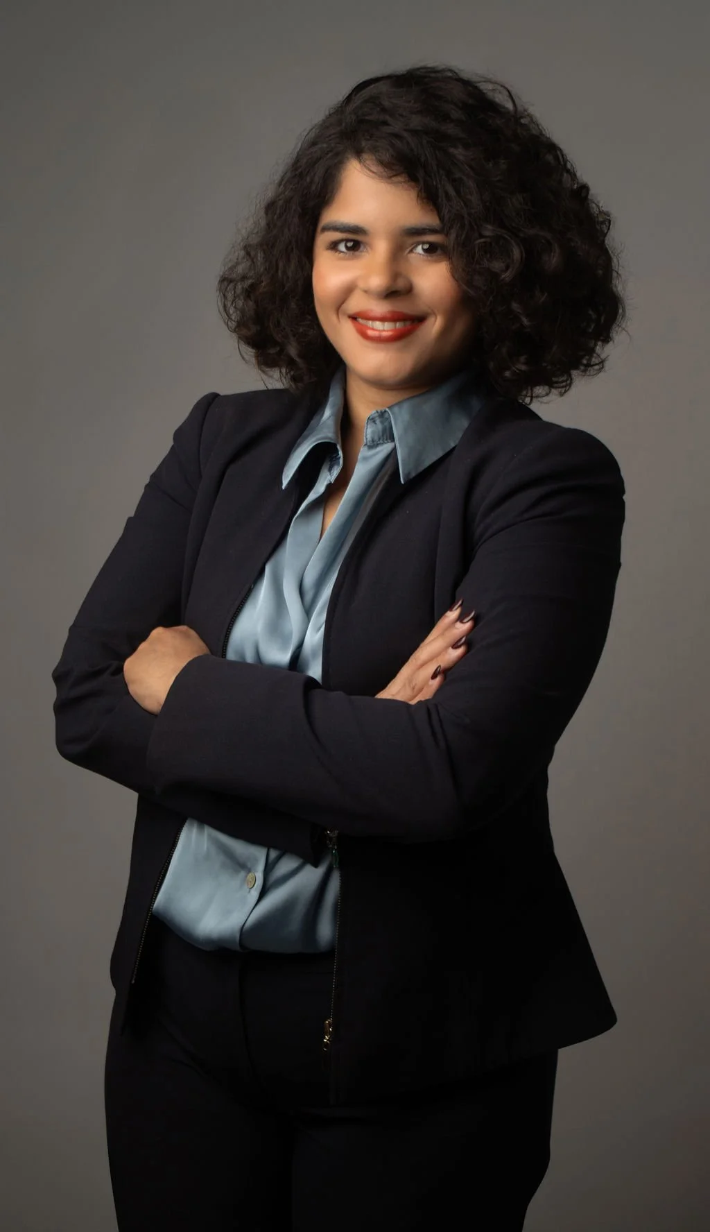 Mujer con cabello rizado, sonriendo, con ropa formal y brazos cruzados, en fondo gris.