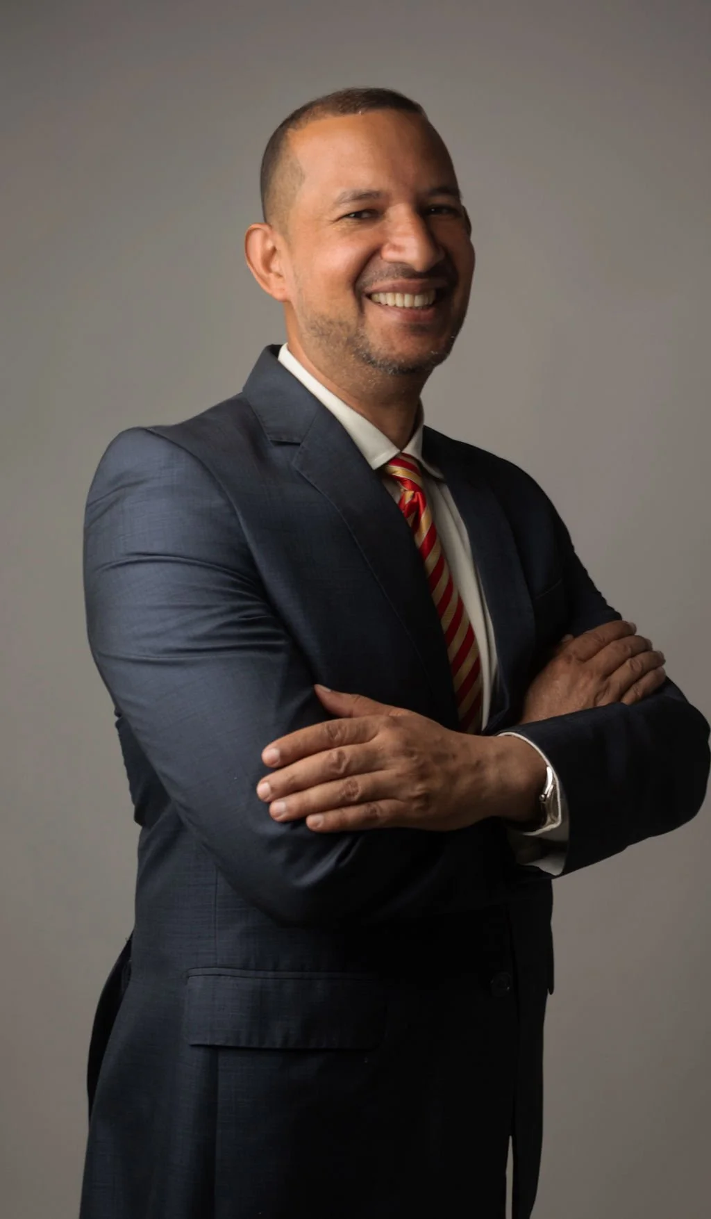 Hombre sonriente con traje oscuro y corbata a rayas, erguido con brazos cruzados frente a un fondo gris.