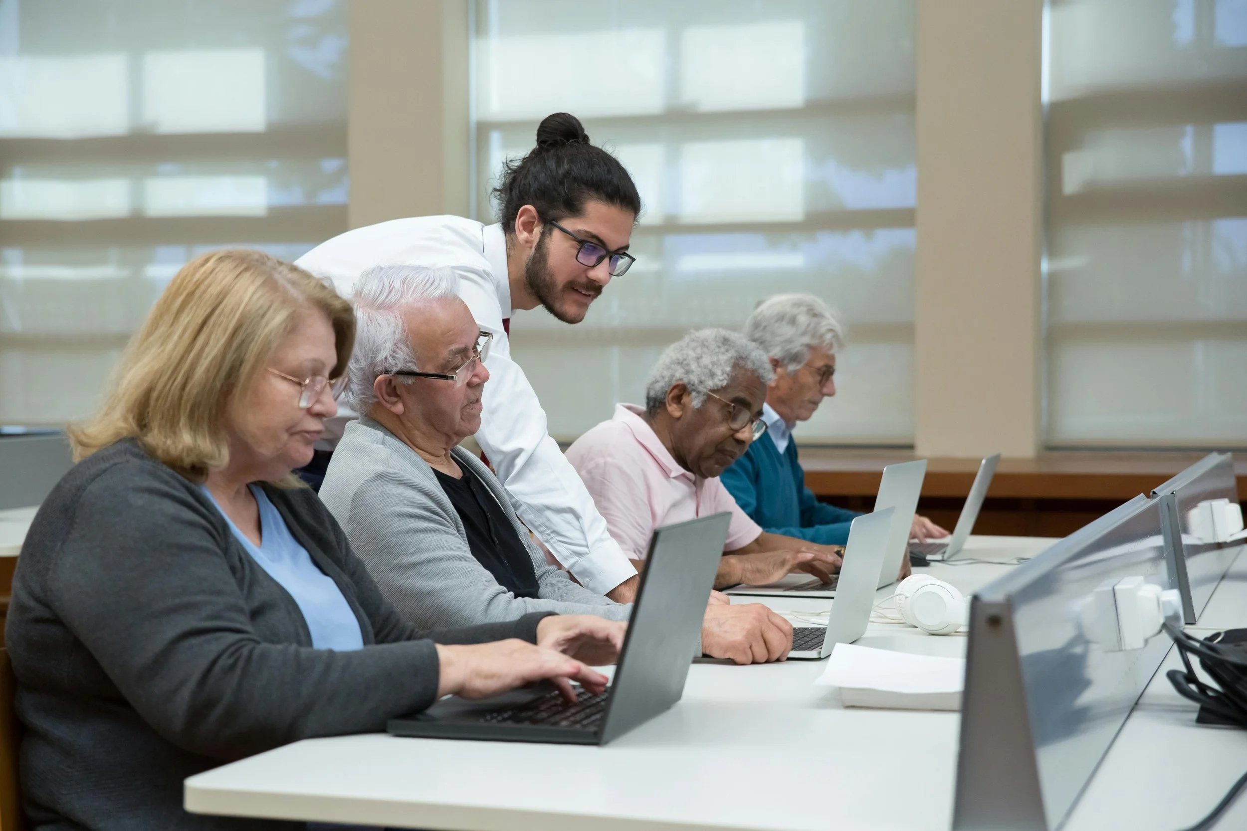 Un grupo de personas mayores sentadas en una mesa, usando computadoras portátiles, mientras un joven les ayuda en un entorno de oficina o aula.