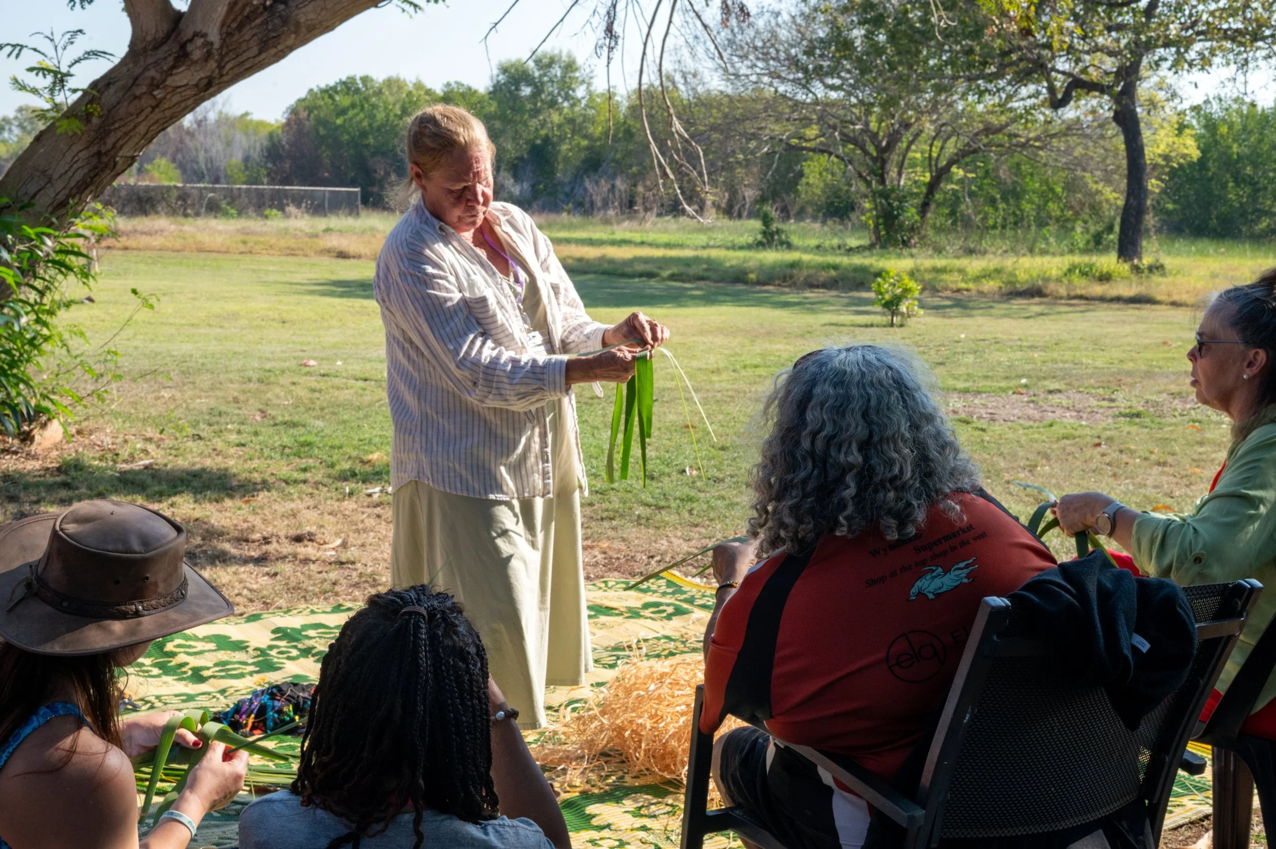 A woman standing outdoors demonstrating weaving or basket-making to a seated group of indigenous women.