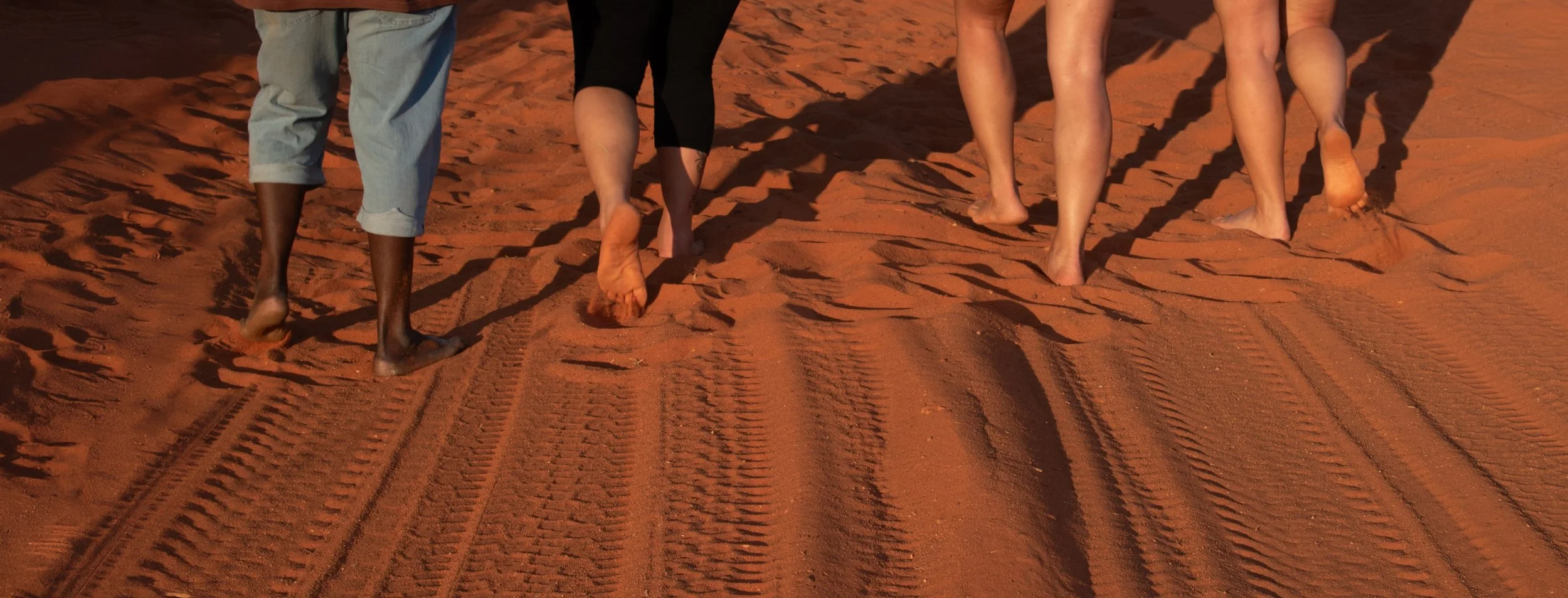 Four people walking barefoot on red sand, leaving footprints behind as they walk up a dune.
