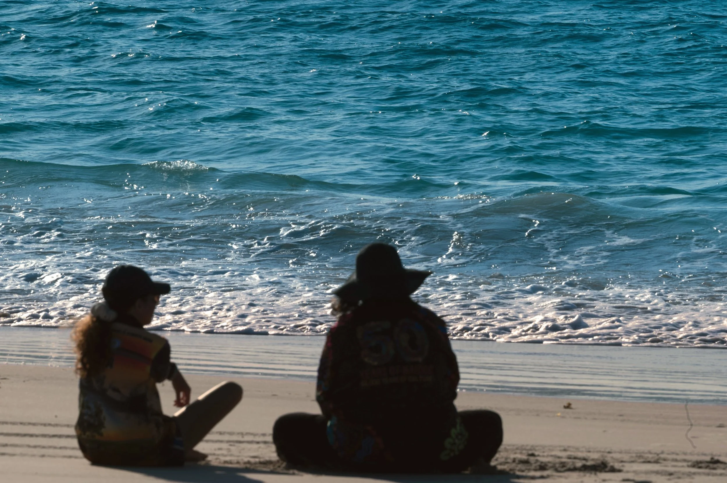 Two indigenous women sitting on the beach, facing the ocean, at community event.