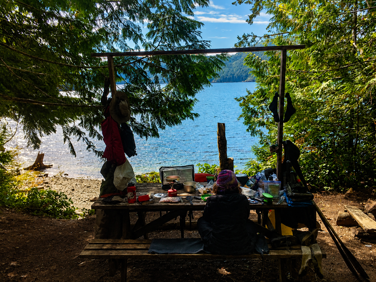 A woman sitting at a picinc table, loaded with gear, at a lakes edge
