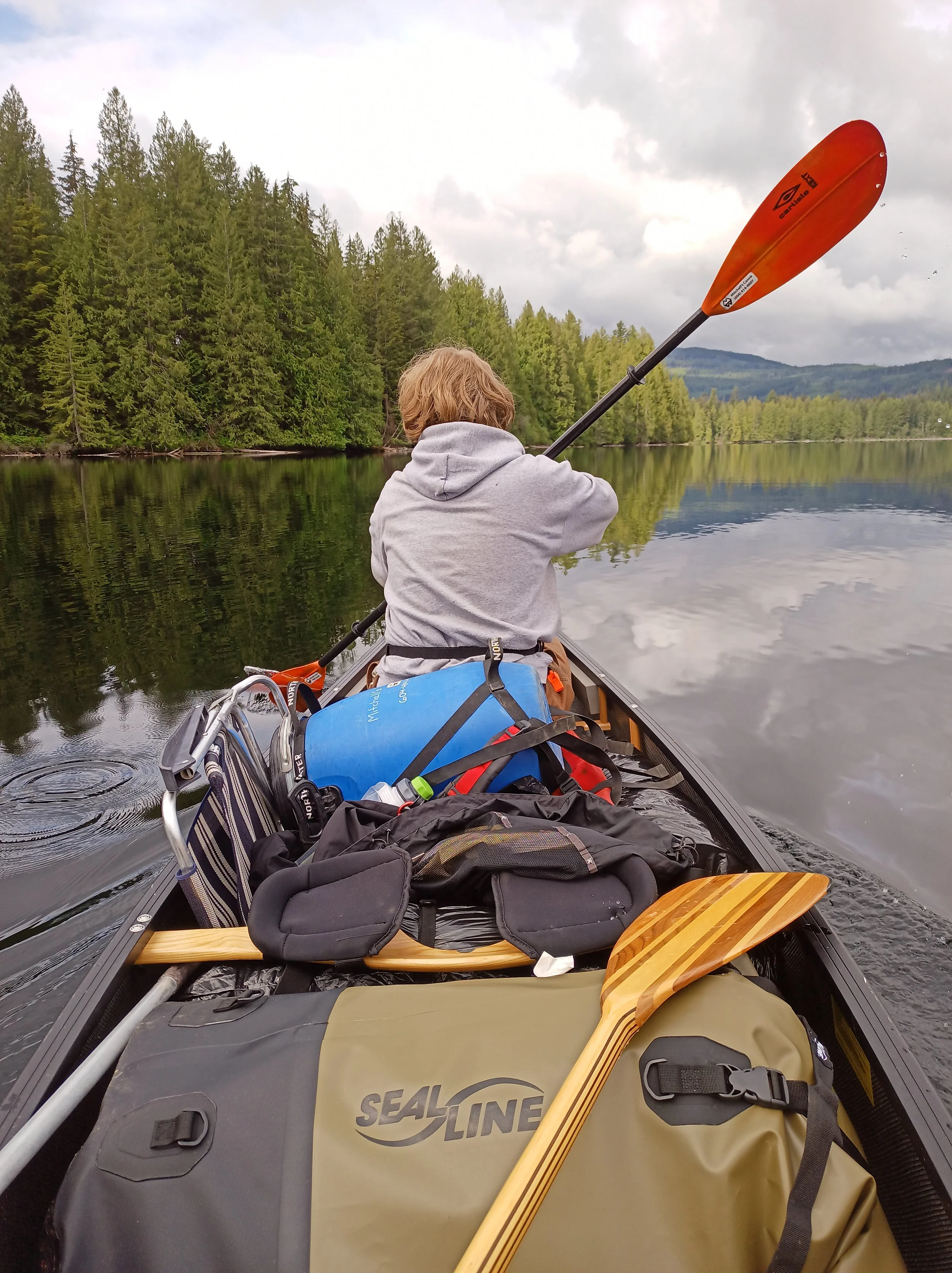 A boy using a kayak paddle in the bow of a canoe loaded with gear.
