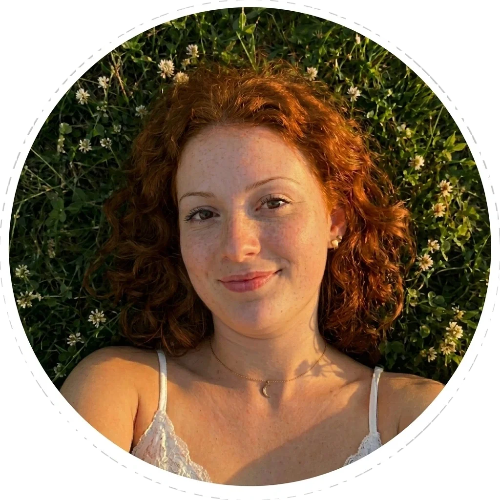 A woman with curly red hair lying on grass with small white flowers, wearing a white lace top and a gold necklace with a crescent moon pendant, smiling at the camera.