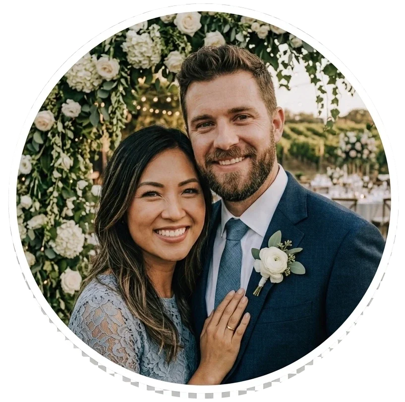 A smiling couple at a wedding, standing close together outdoors under a floral arch, with scenic greenery and string lights in the background.