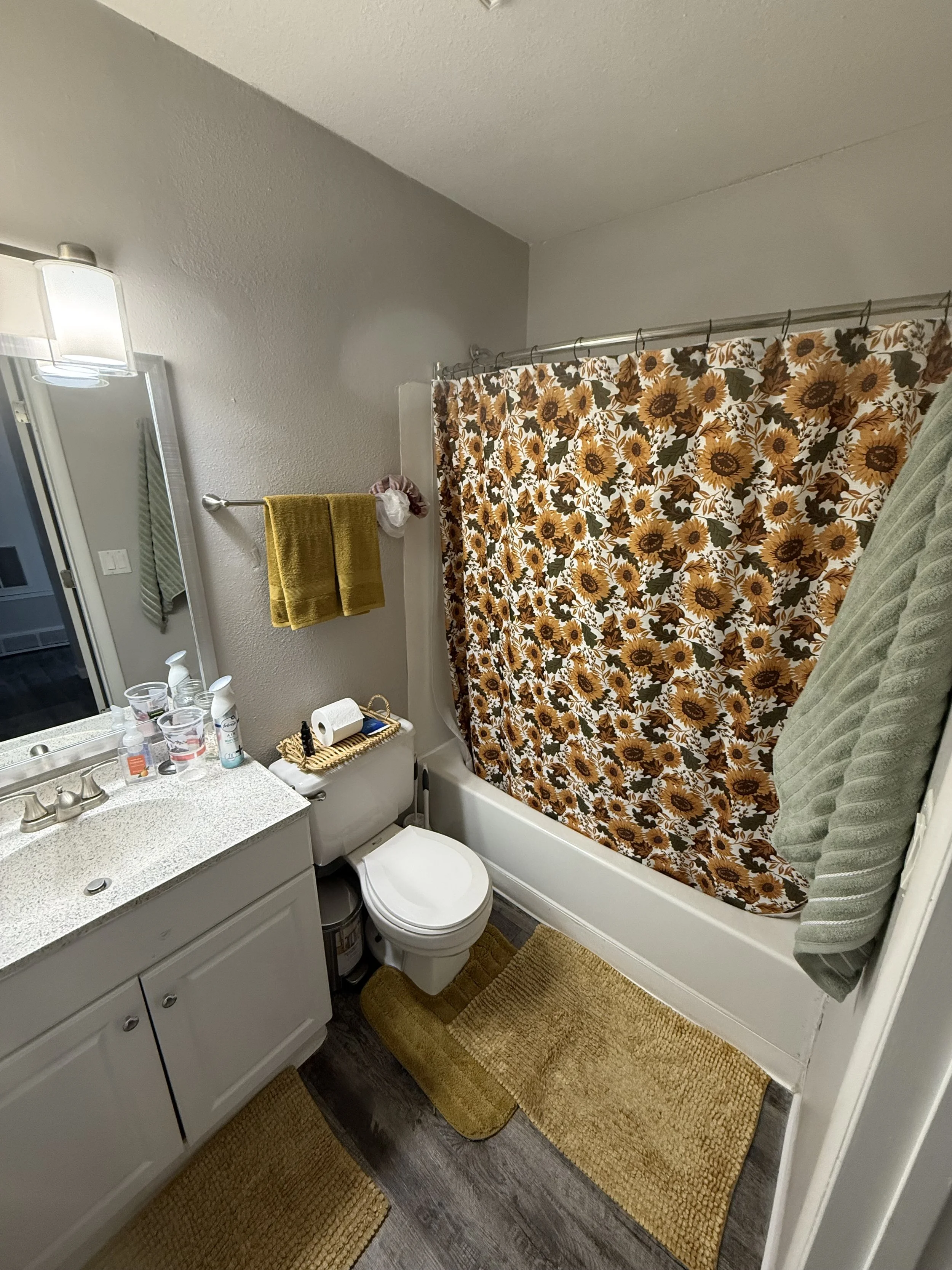 A bathroom with a white vanity, a sink with soap bottles, a small mirror, a toilet, a yellow rug, a towel rack with yellow towels, a shower with a sunflower-patterned curtain, and a green towel hanging on the side.
