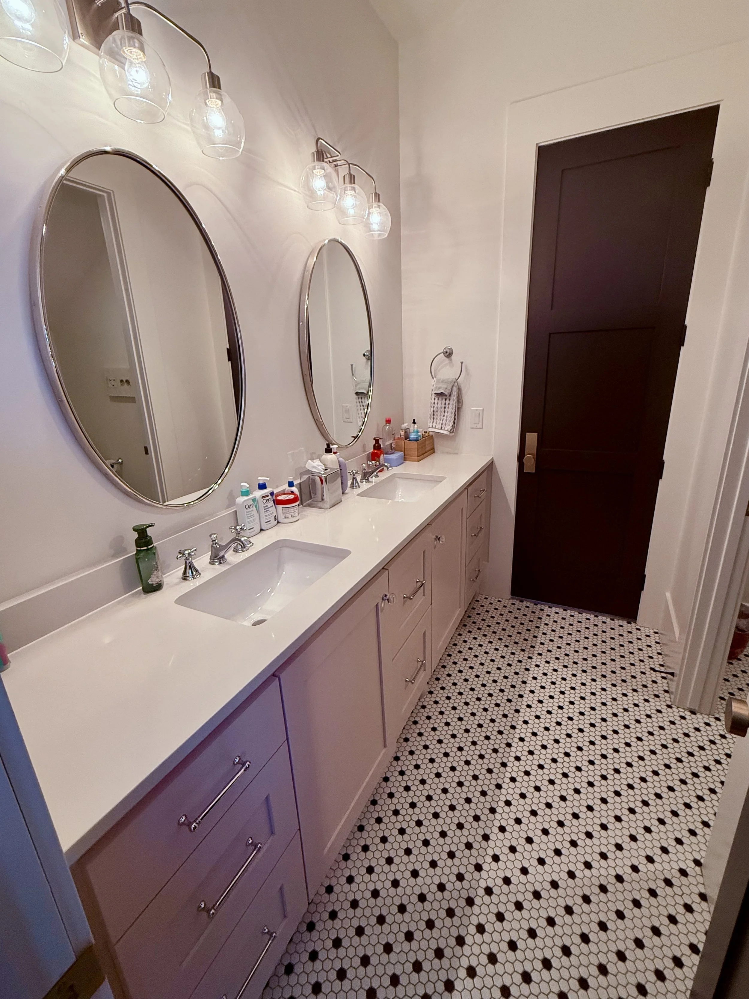 A bathroom with a light-colored vanity, oval mirrors, and patterned black-and-white tile floor.