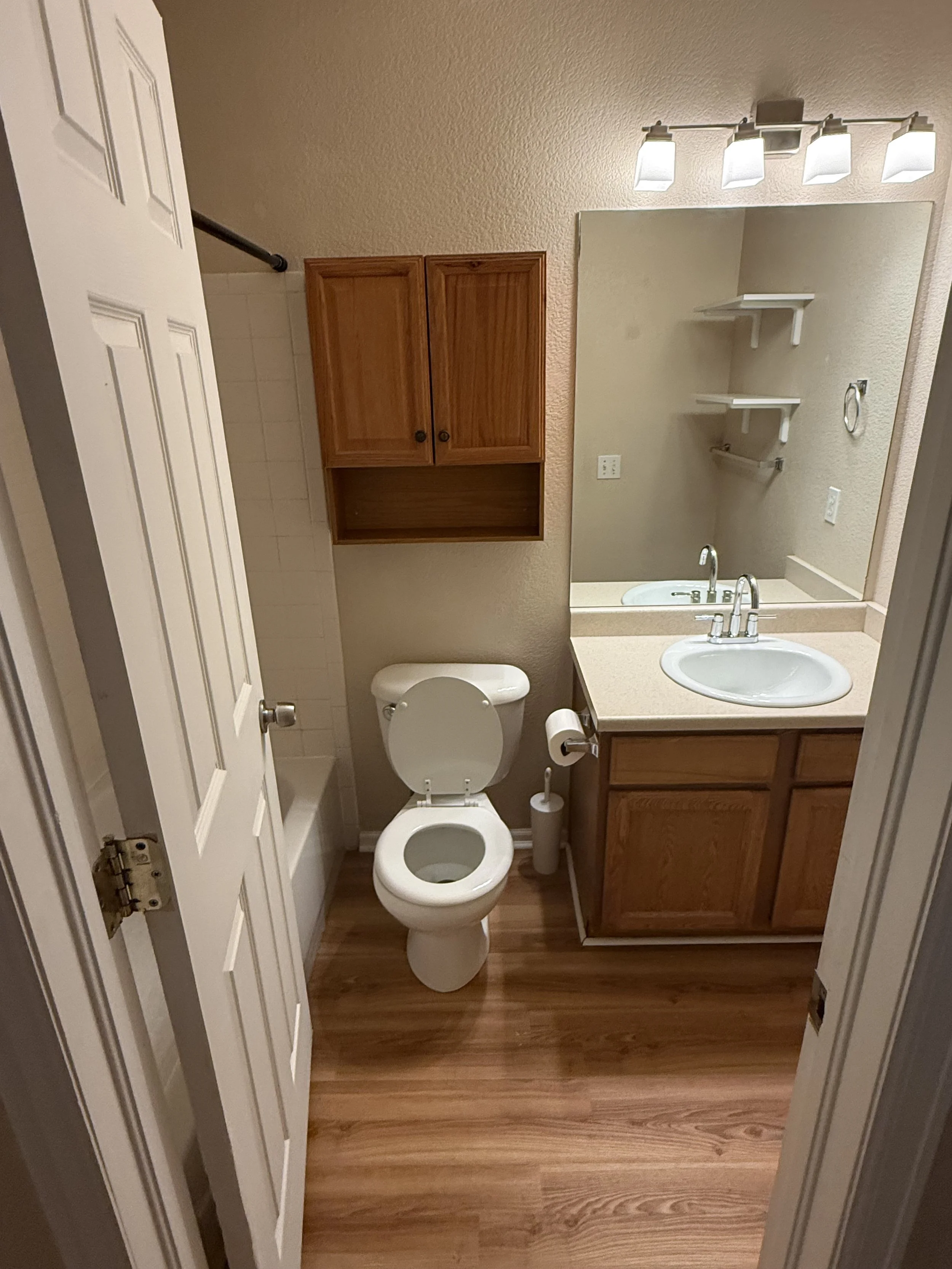 Small bathroom with a toilet, wooden cabinet above, mirror, sink, and shelves. The floor is wood, and the walls are painted beige.