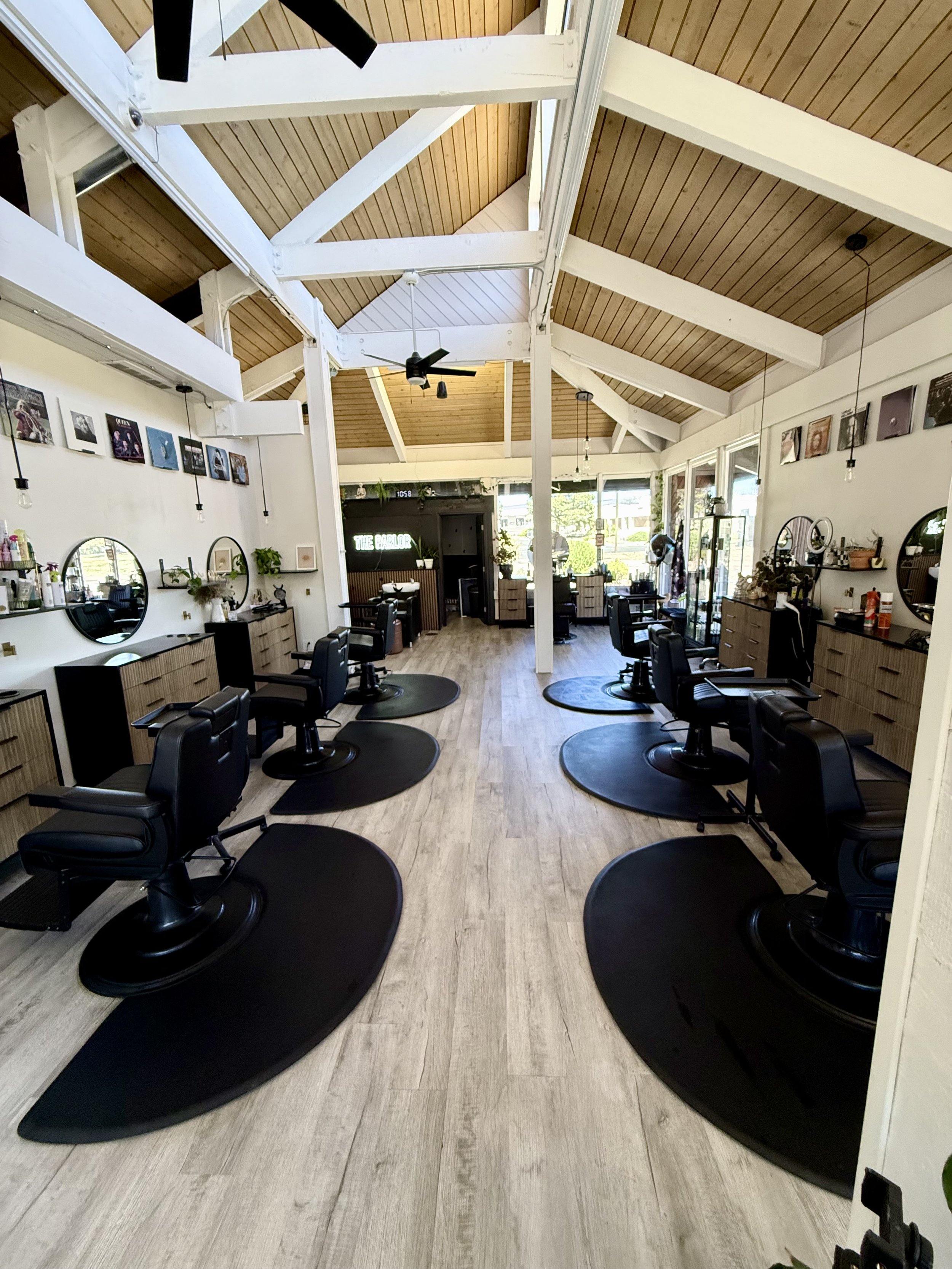 Interior of a modern hair salon with black styling chairs, large mirrors, and salon supplies, featuring a wooden ceiling with white beams and natural light from large windows.