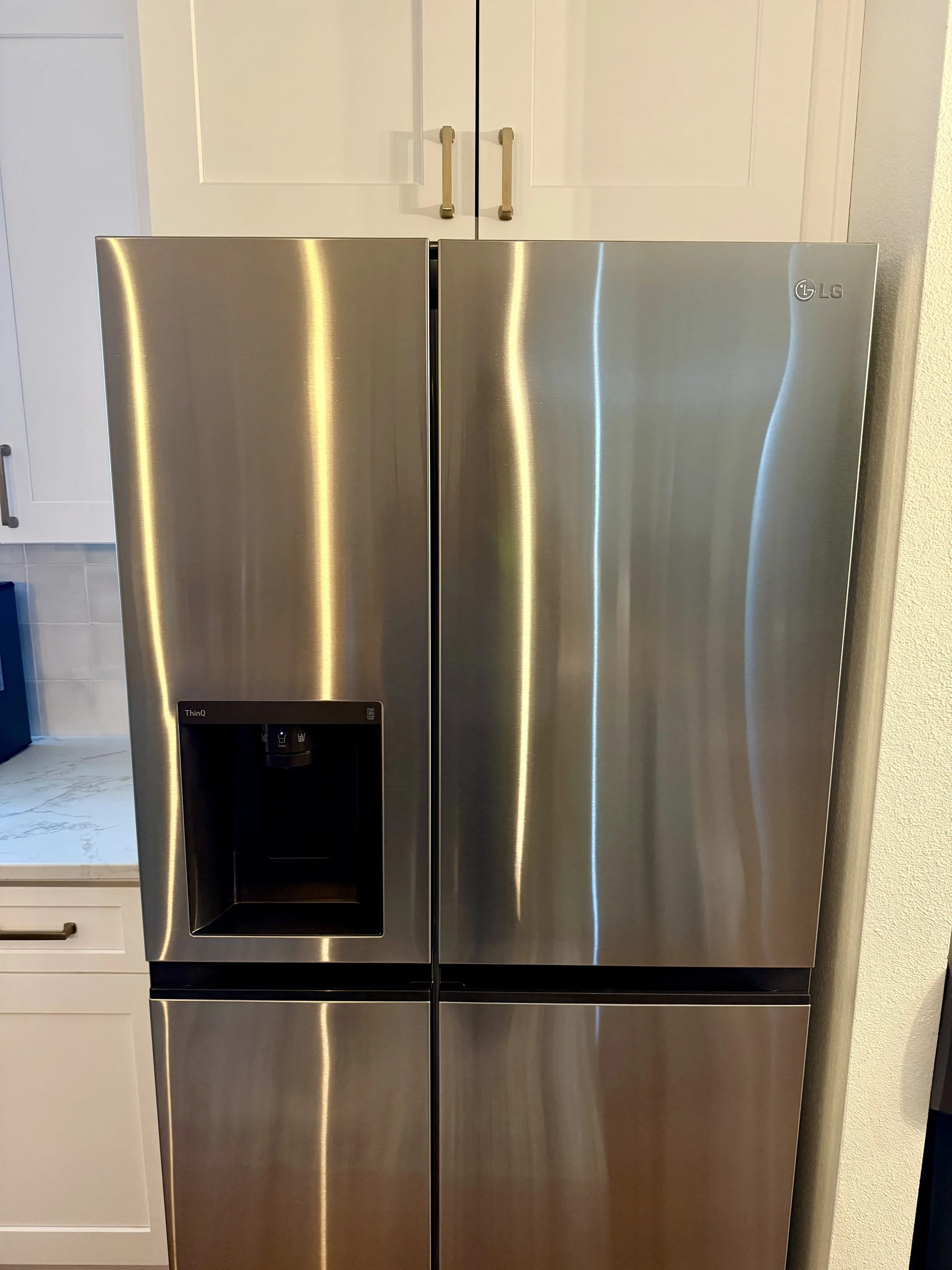 Stainless steel LG side-by-side refrigerator with water and ice dispenser, situated in a kitchen with white cabinets and light-colored walls.