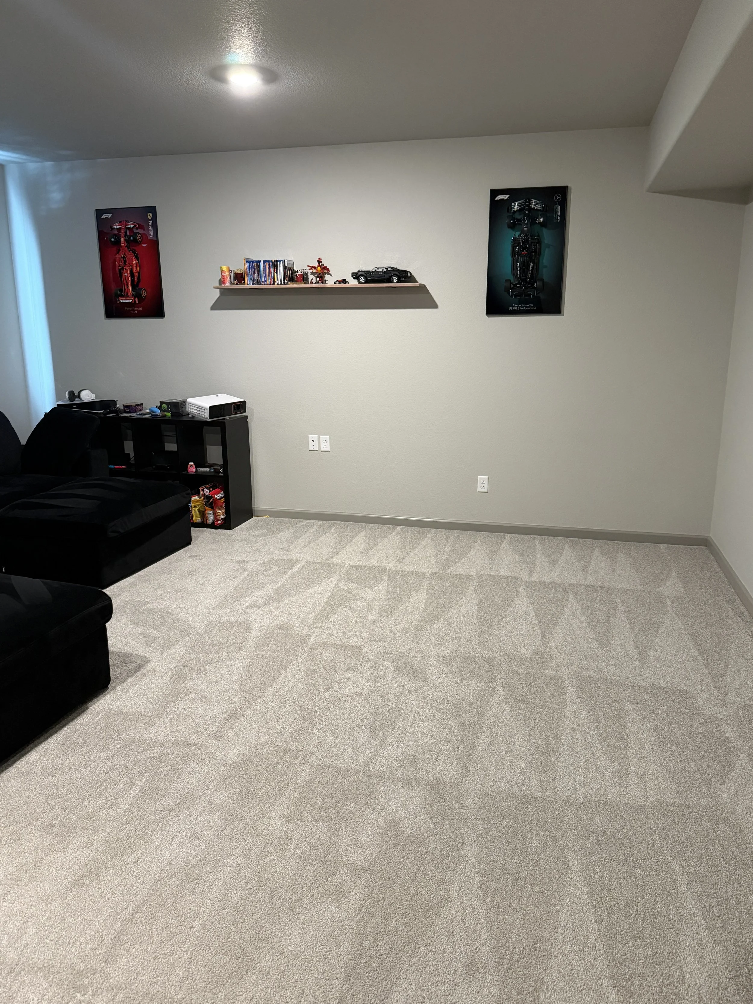 A basement room with beige carpet, a white wall, and two framed posters of Formula 1 cars on the wall.