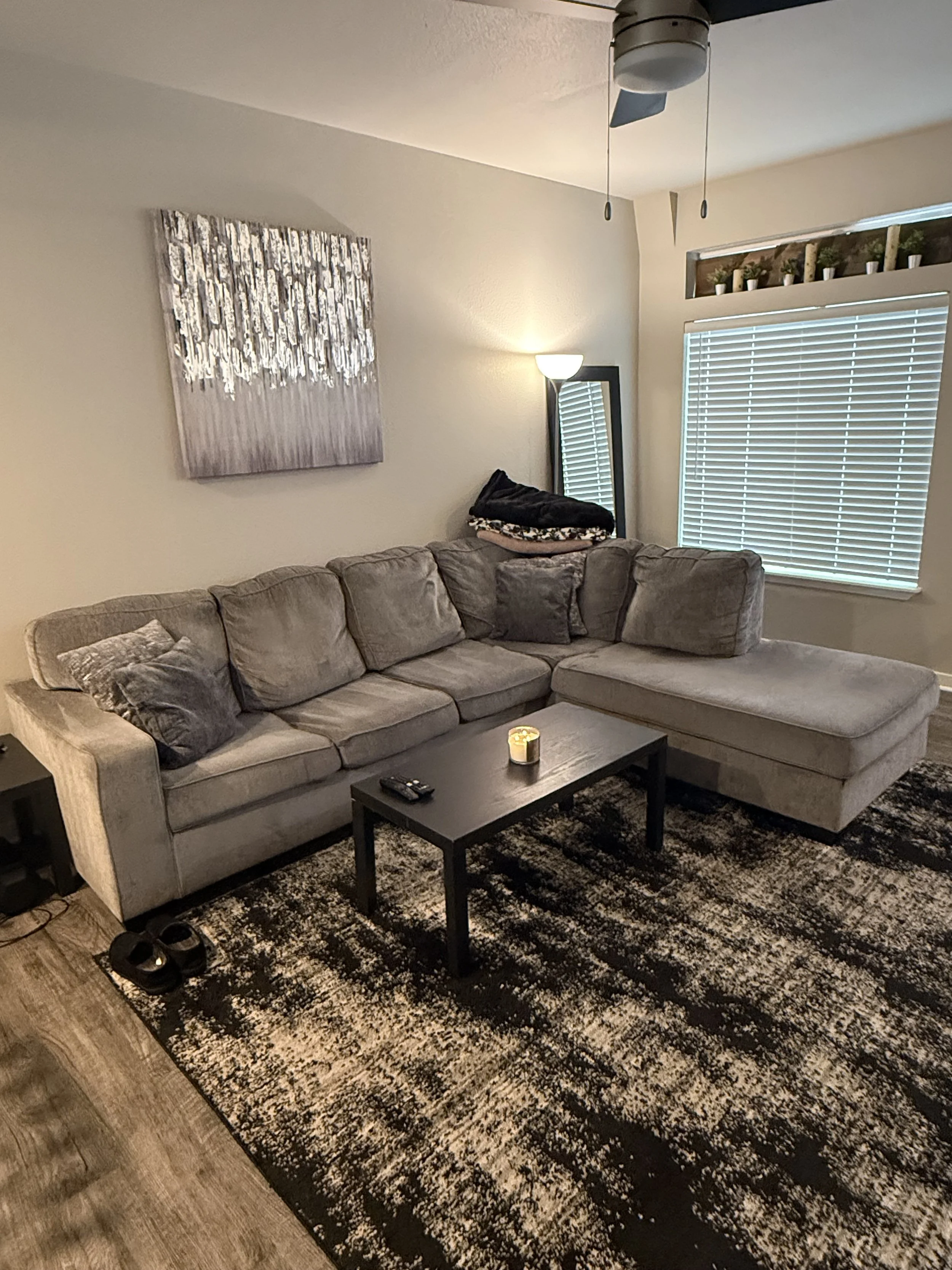 Living room with beige sofa, black coffee table, area rug, window with blinds, wall mirror, wall art, ceiling fan, and small candles on the table.