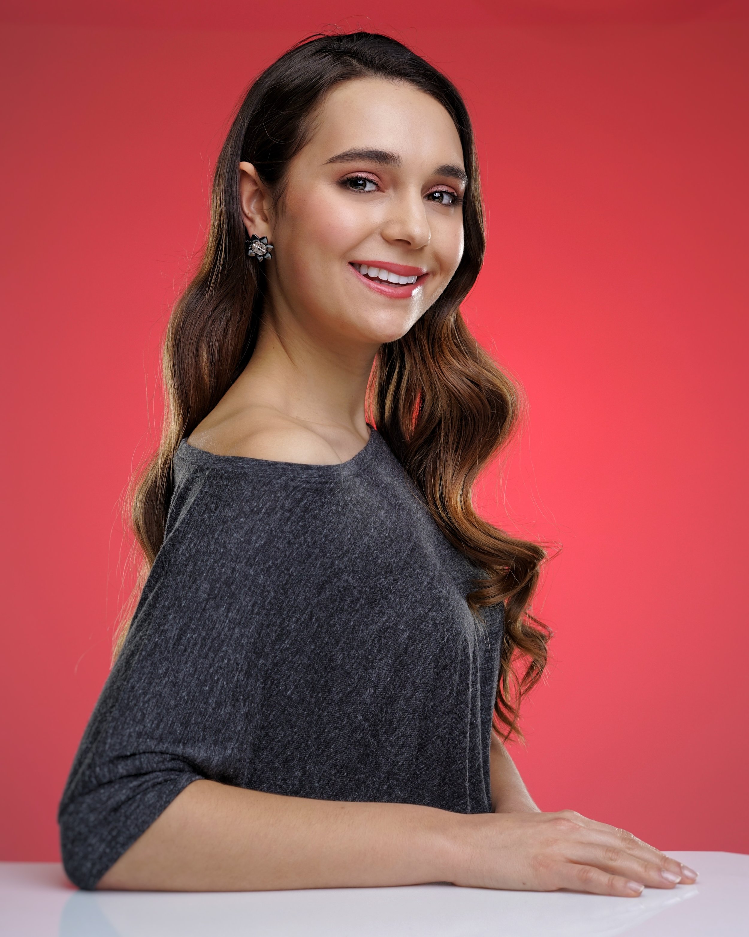 A woman with long brown hair wearing a gray off-shoulder top poses against a red background, smiling, with one hand resting on a table.