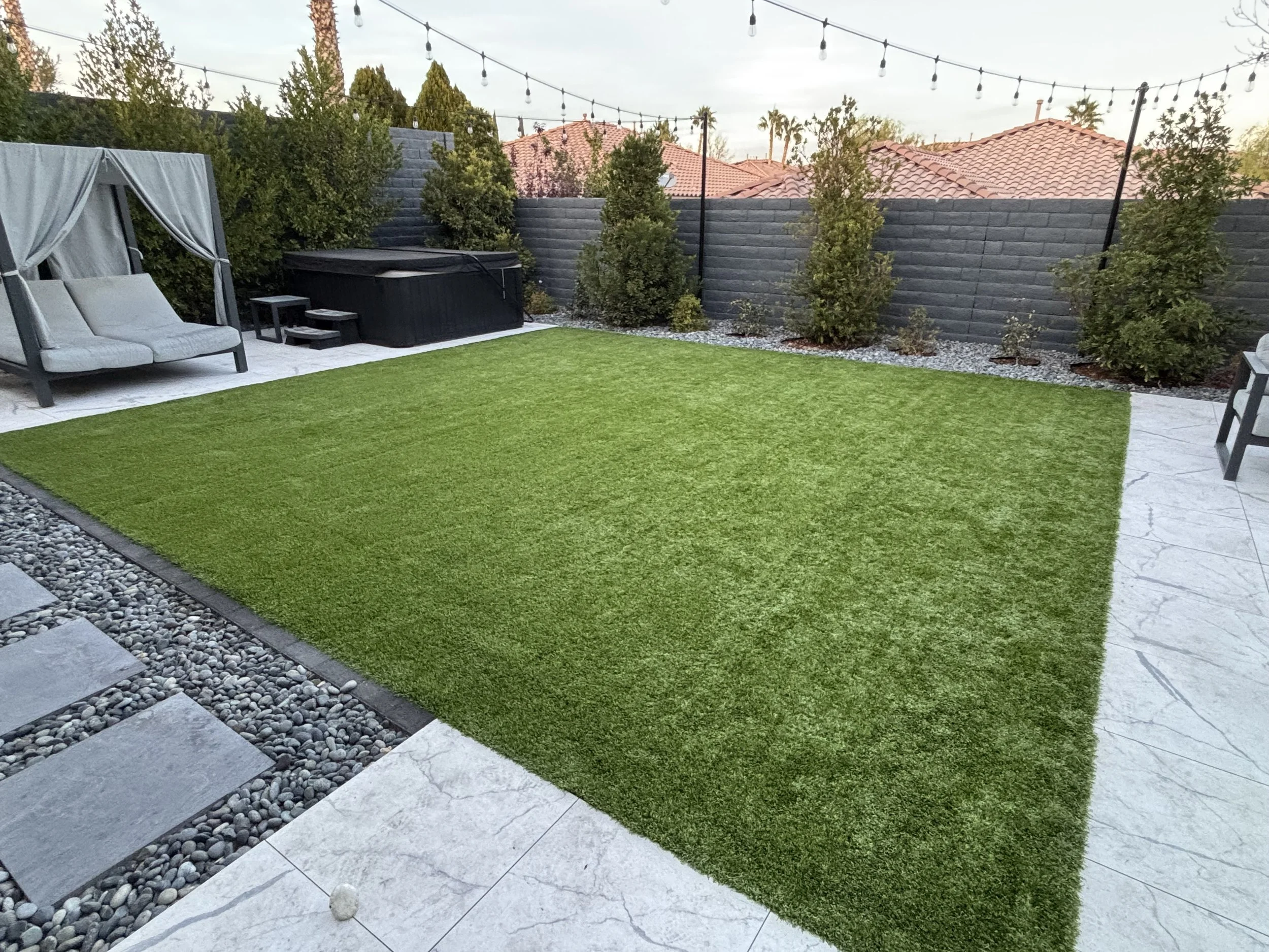 A backyard patio with a green artificial grass lawn, surrounded by a gray stone border, with seating, a hot tub, and string lights overhead.