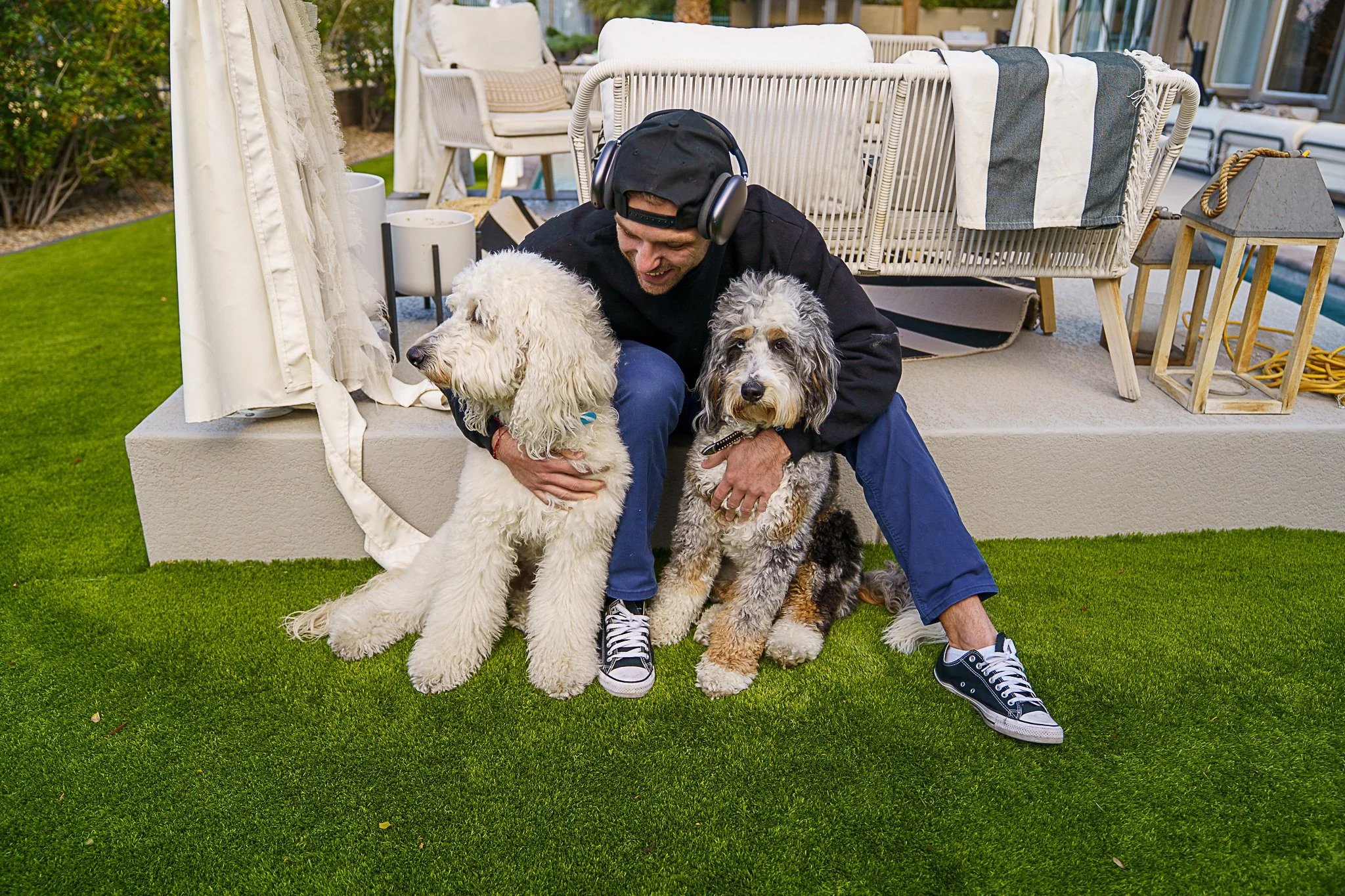 A man with headphones crouches on the grass, holding two dogs, one white and one gray, in a backyard with outdoor furniture and a green lawn.