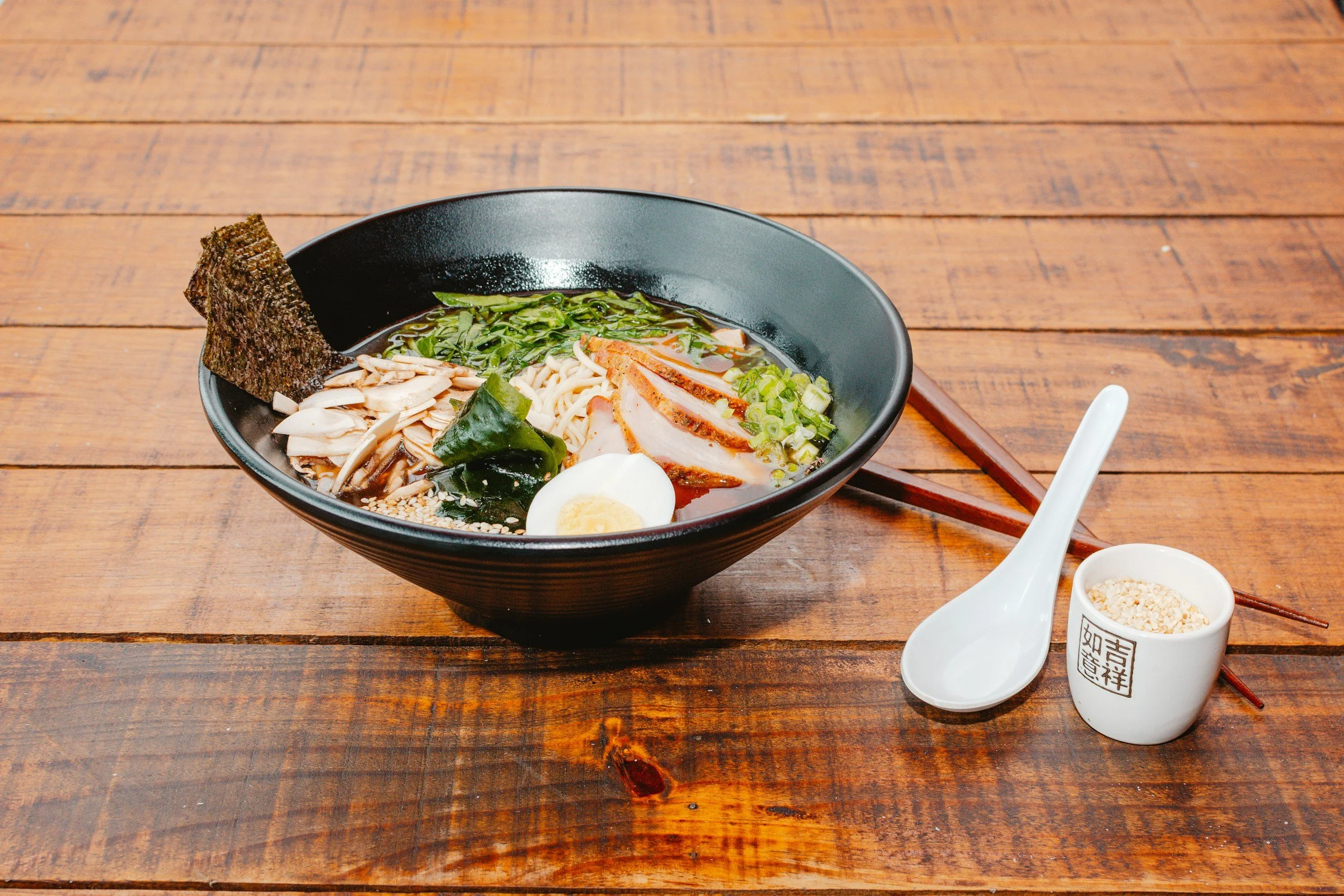 Ramen bowl with sliced meat, seaweed, boiled egg, mushrooms, green onions, and broth, on a wooden table with chopsticks and a spoon.
