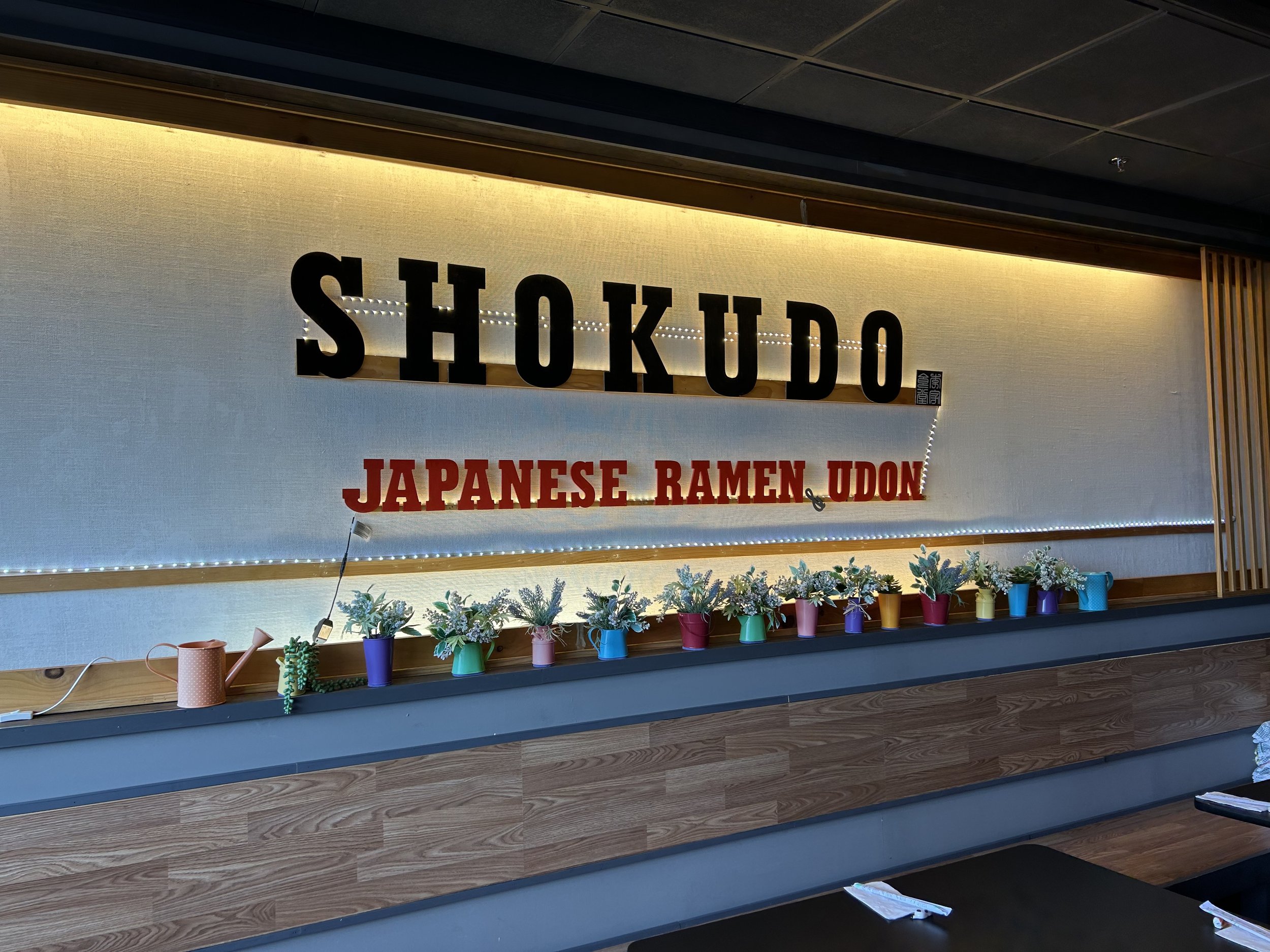 Japanese restaurant interior sign reading 'Shokudo Japanese Ramen Udon' with decorative plants on a shelf below.