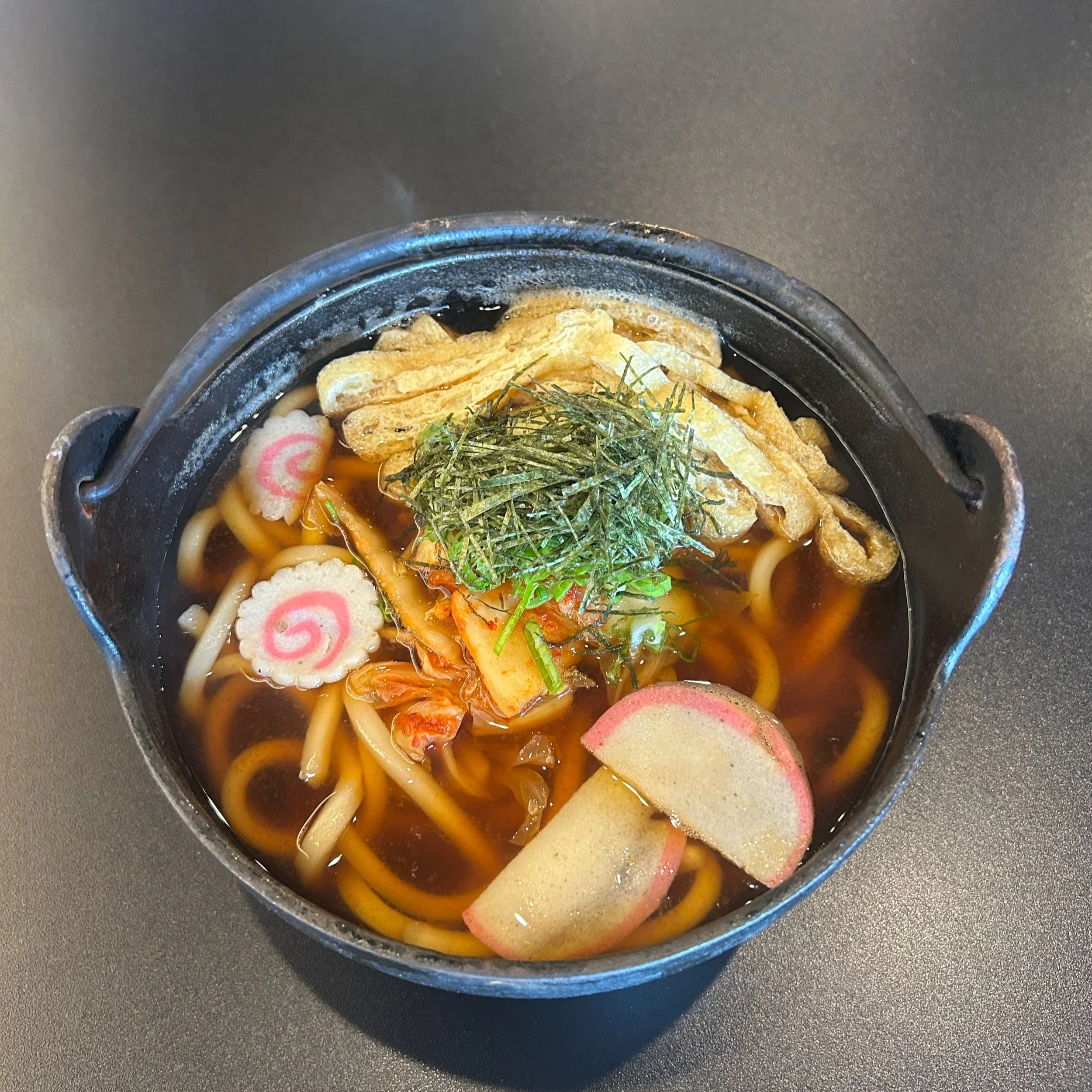 A bowl of Japanese udon soup with thick noodles, narutomaki slices, tempura bits, seaweed, and green onions in a dark broth.