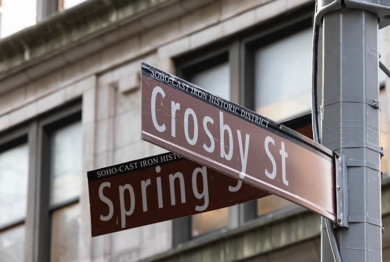 Street signs for Crosby Street and Spring Street in the Soho-Cast Iron Historic District, Manhattan, New York City, showing part of a building with large windows in the background.