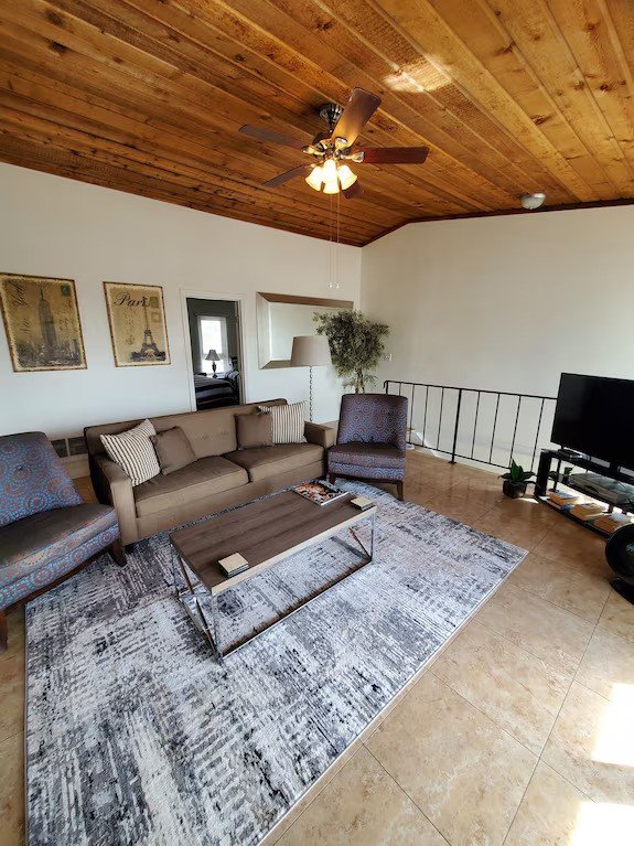 Living room with a beige sofa, two patterned chairs, a wooden coffee table on a gray area rug, a TV, wooden ceiling with fan, and framed Paris-themed art on the wall.