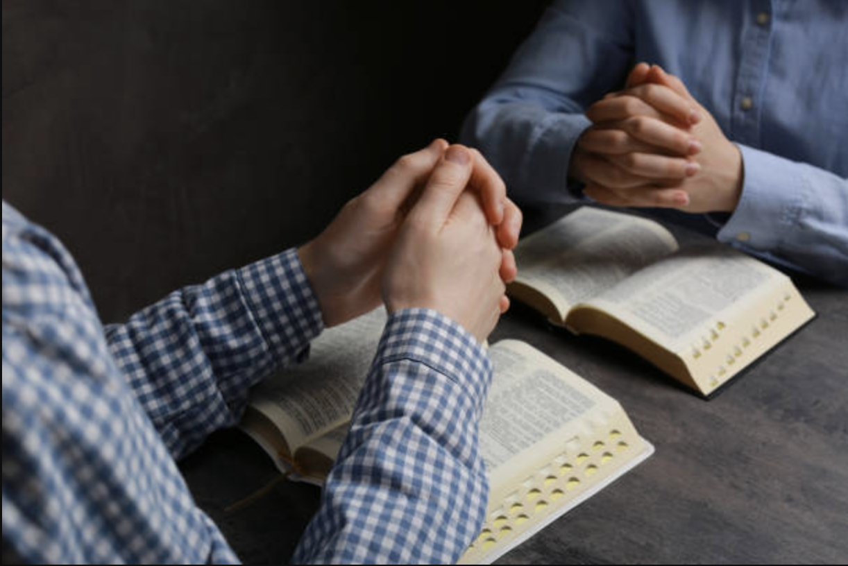 Two people are sitting at a table with open Bibles, clasping their hands in prayer.