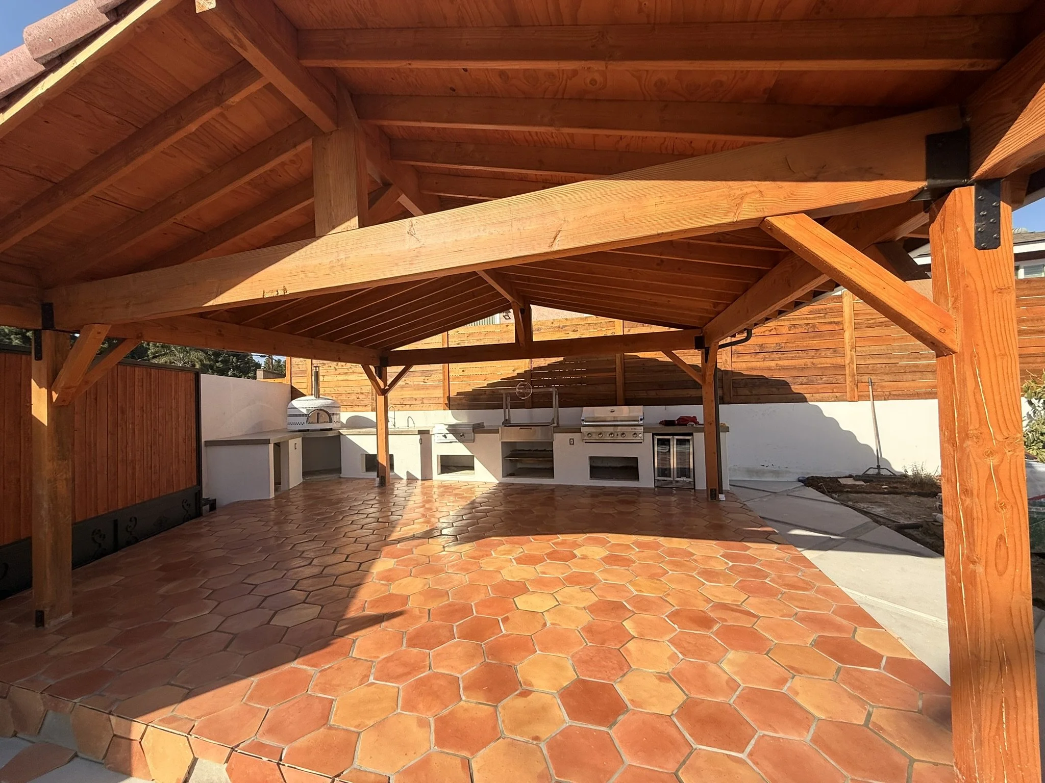 PAVERS & OUTDOOR LIVING | Outdoor kitchen beneath a timber pavilion, with grill, counters, and tile flooring.
